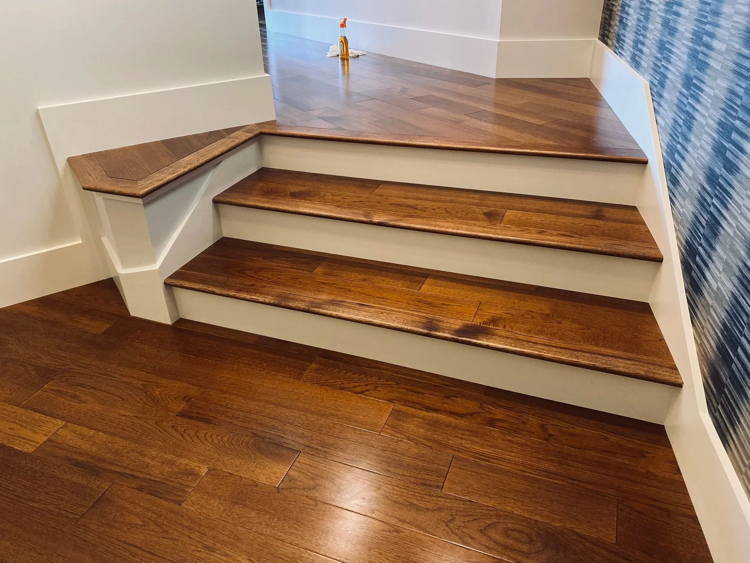 Wooden staircase with three steps and a hardwood floor, adjacent to a wall with blue textured wallpaper and a white trim.