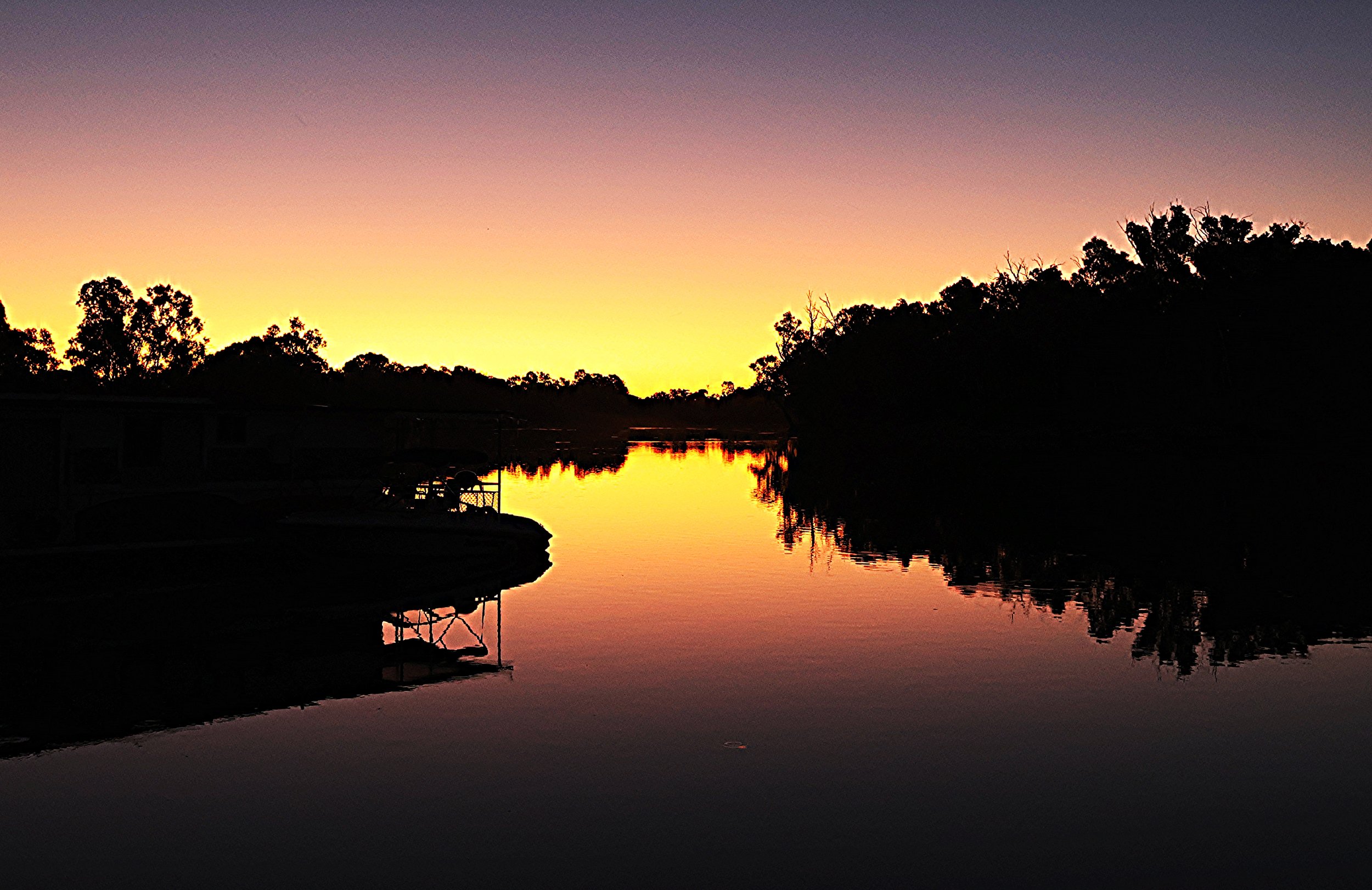 A river at sunset with the sky glowing orange and purple, silhouetted trees on either side, and their reflections in the water.