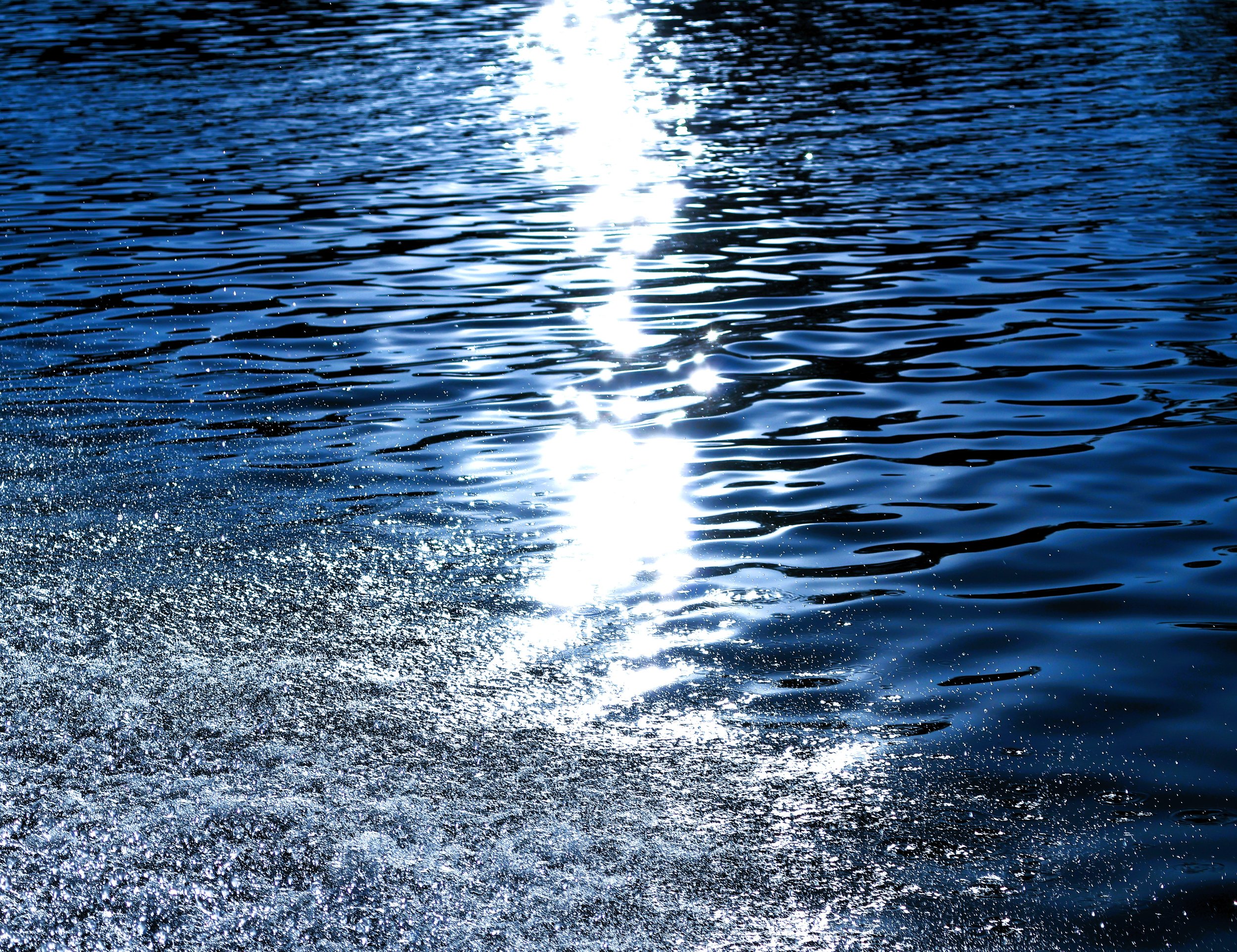 Sunlight reflecting on blue water with ripples, and foam at the bottom left corner.