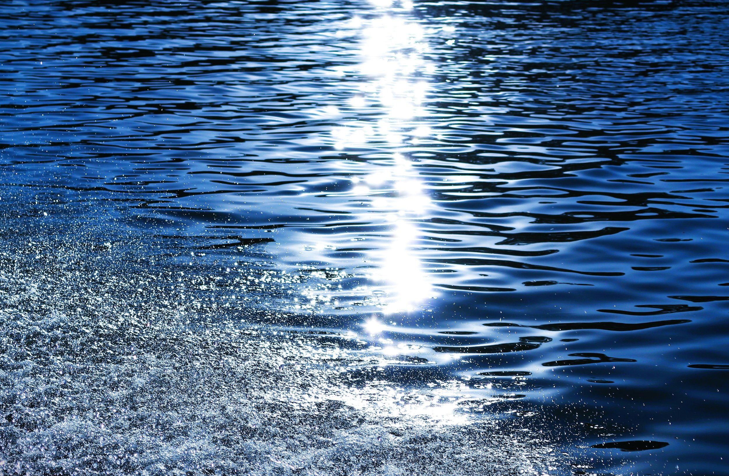 Sunlight reflecting on rippling blue water, with trail of foam and droplets at the bottom left.