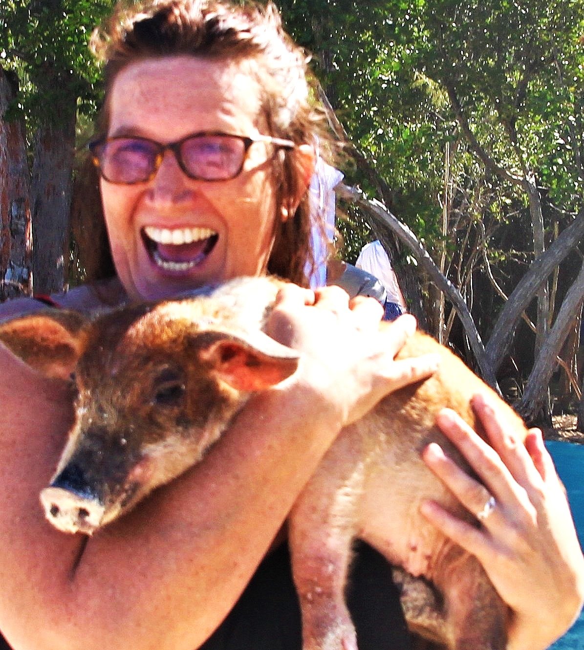 A woman wearing glasses holding a piglet outdoors, smiling and laughing with her mouth open. There are trees and greenery in the background.