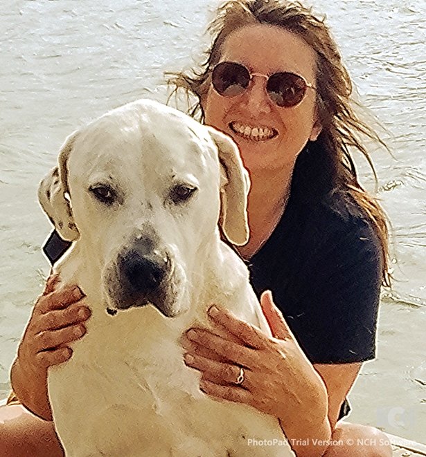 A woman wearing sunglasses smiling and holding a large white dog near a body of water.