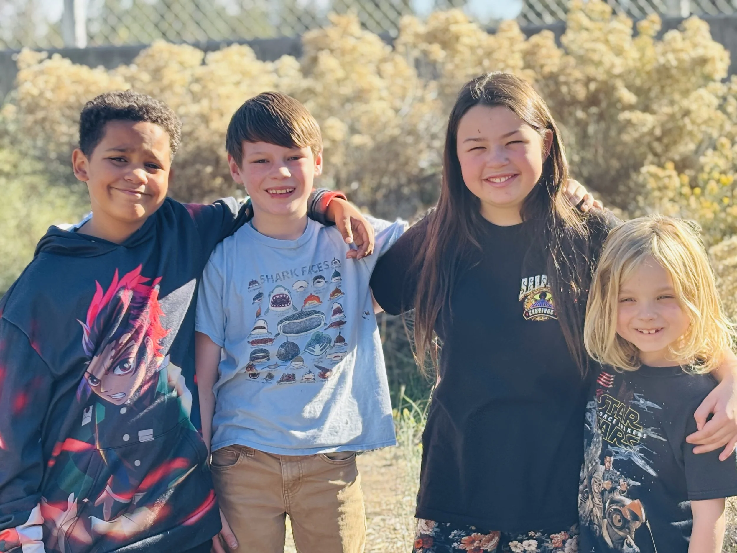 Four children standing outdoors, smiling, with trees and bushes in the background.