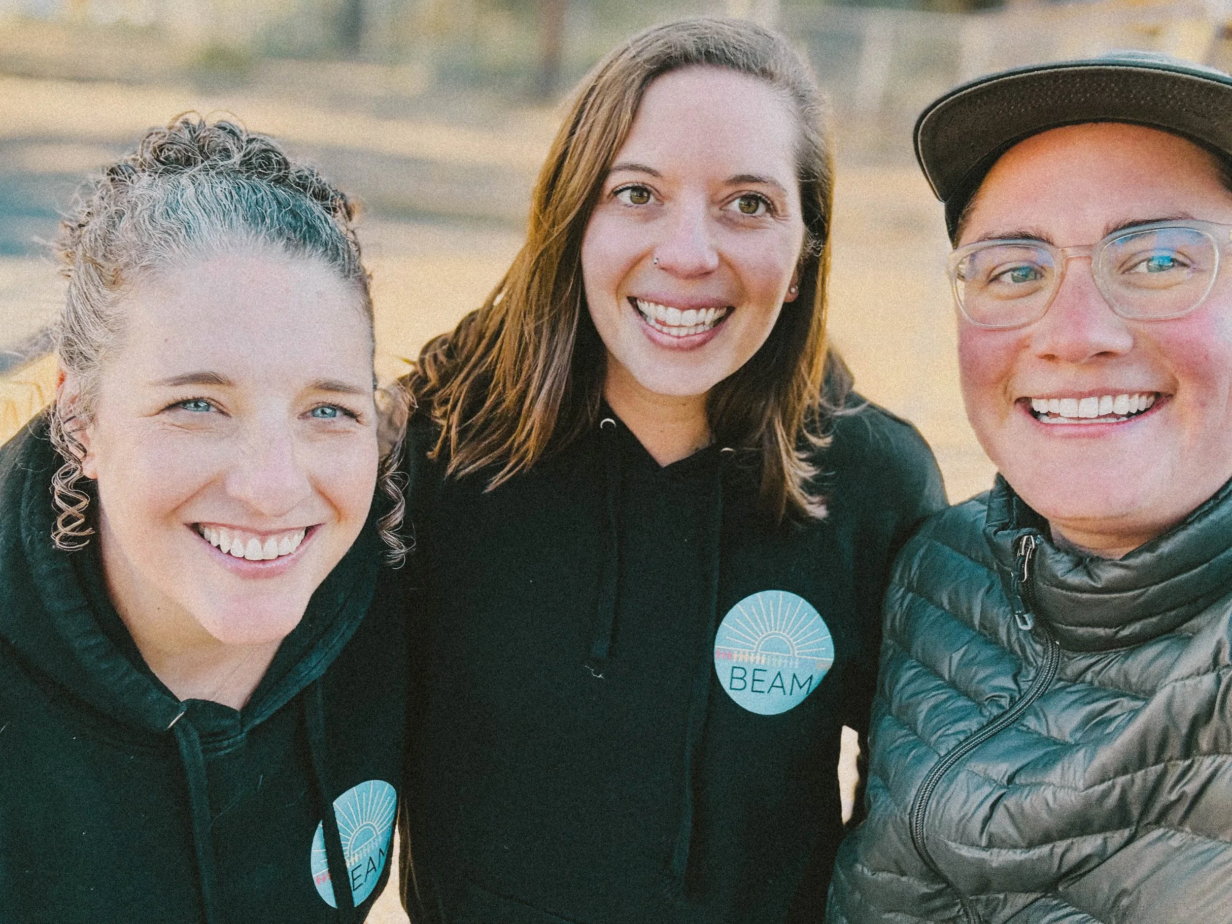 Three smiling people outdoors, wearing jackets, with a badge that says 'BEAM' on their shirts.