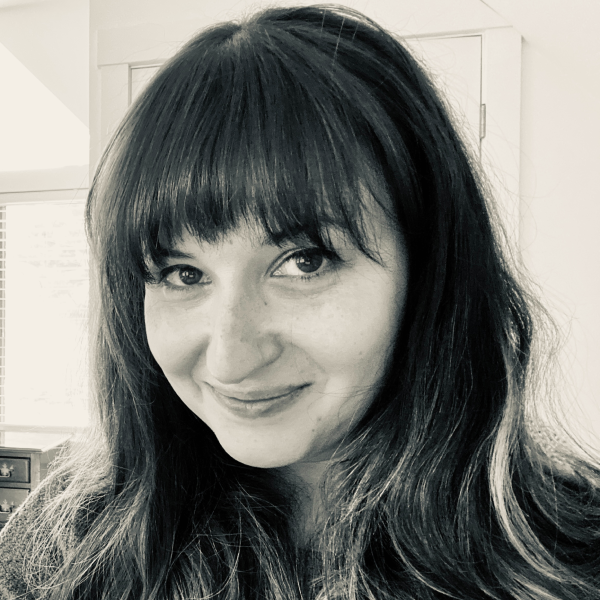 A woman with long dark hair and bangs, smiling at the camera, in a black and white photo with a background of a window and a wooden cabinet.