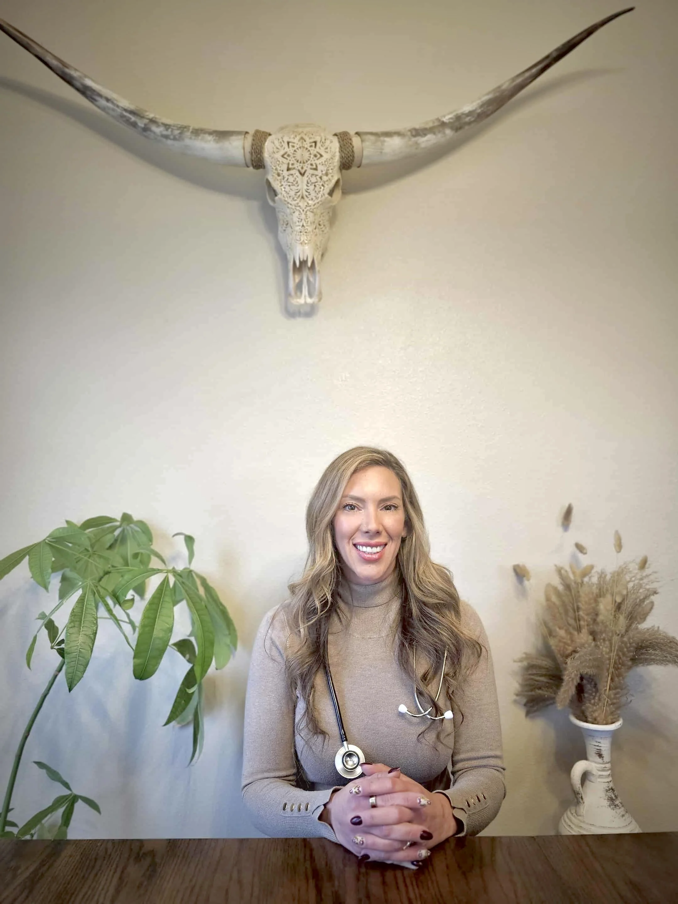 A woman sitting at a wooden table, smiling at the camera, with a stethoscope around her neck. Behind her, there is a large leafy plant on the left and a white vase with dried pampas grass on the right. A decorative longhorn skull is mounted on the wall above her.