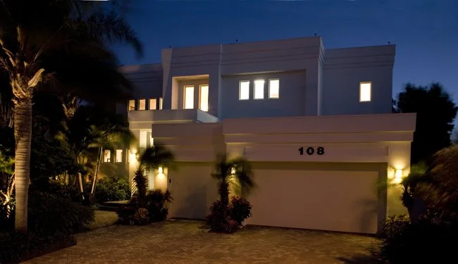 Modern two-story house with illuminated windows and landscaping, seen at night.