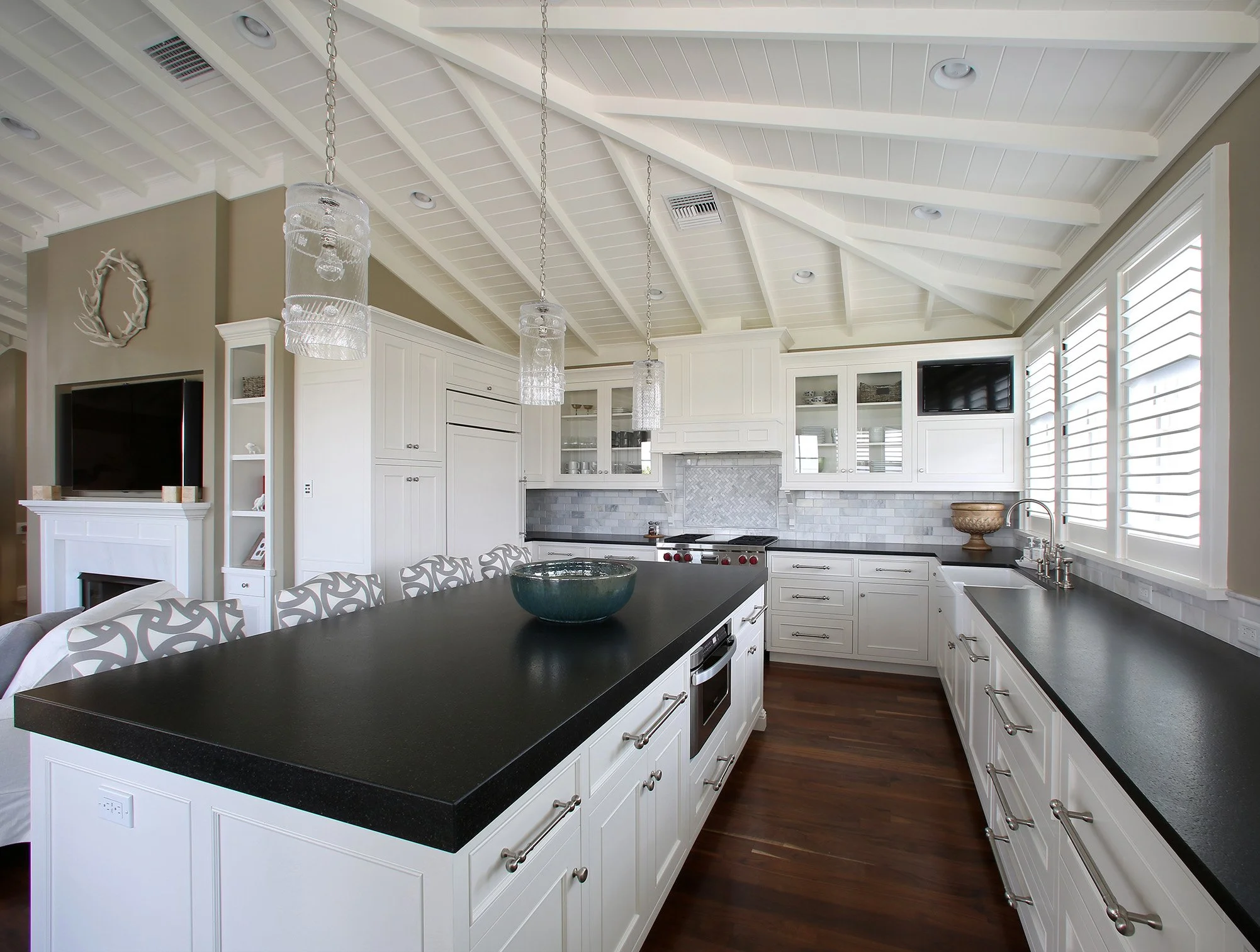 Modern kitchen with white cabinets, black countertops, hanging pendant lights, and a large island, featuring a stove, sink, and window shutters.
