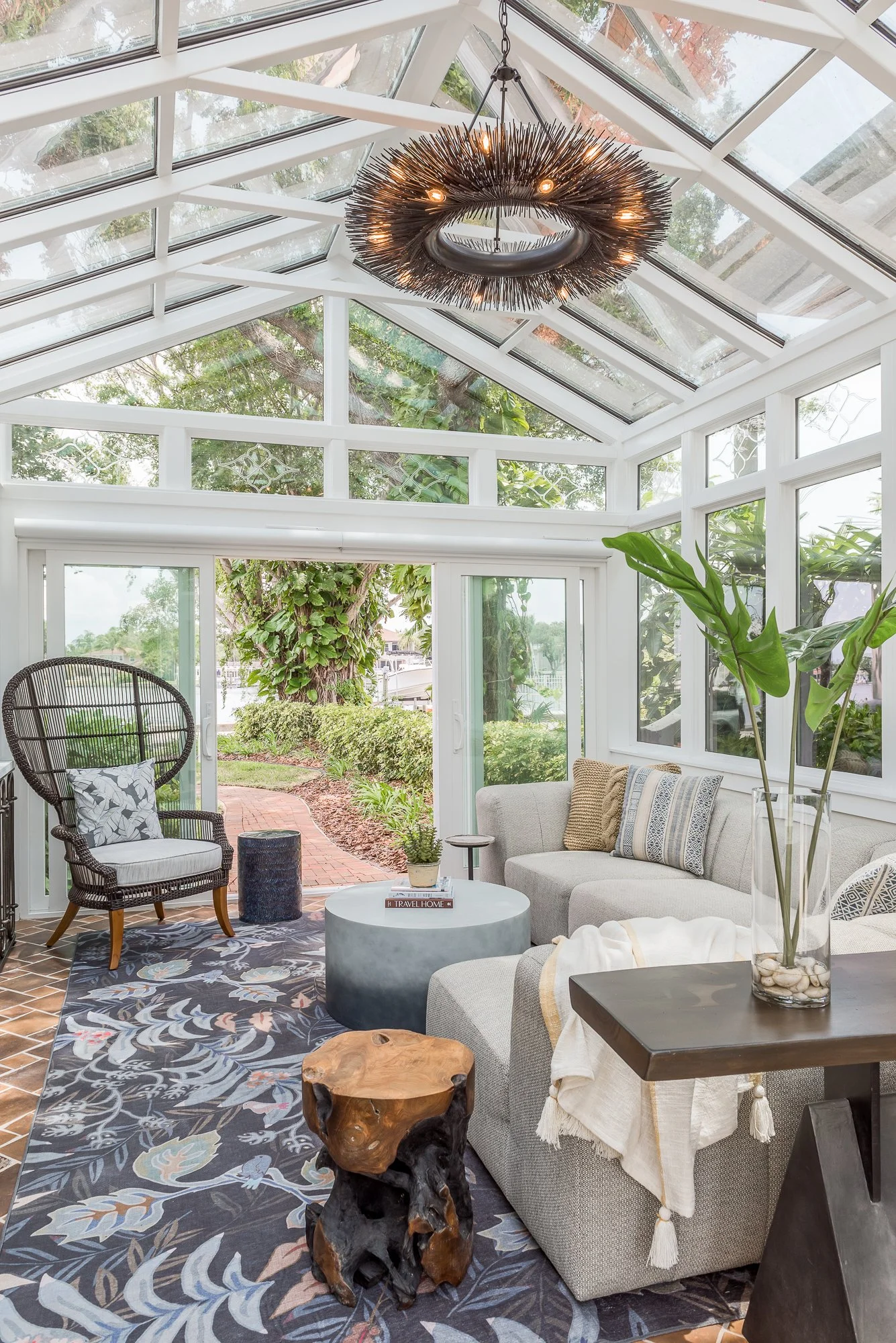 Sunroom with glass walls and ceiling, furnished with a beige sofa, black wicker chair, round white coffee table, wooden side table, and a large potted plant. A decorative ring chandelier hangs from the ceiling, and outside you can see trees and greenery.