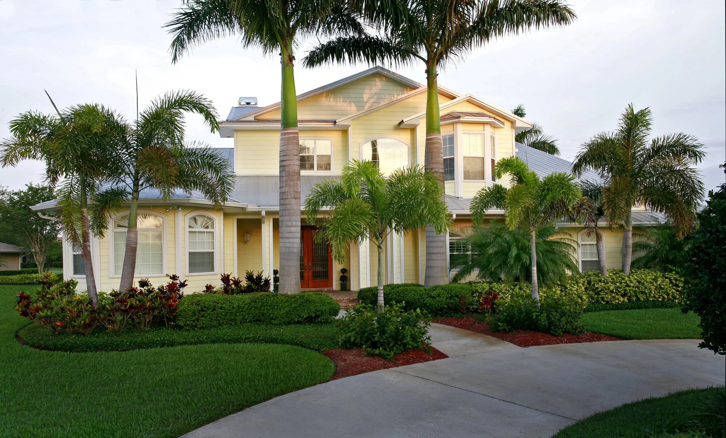 A two-story yellow house with a metal roof, surrounded by lush green landscaping and tall palm trees, with a curved driveway in front.