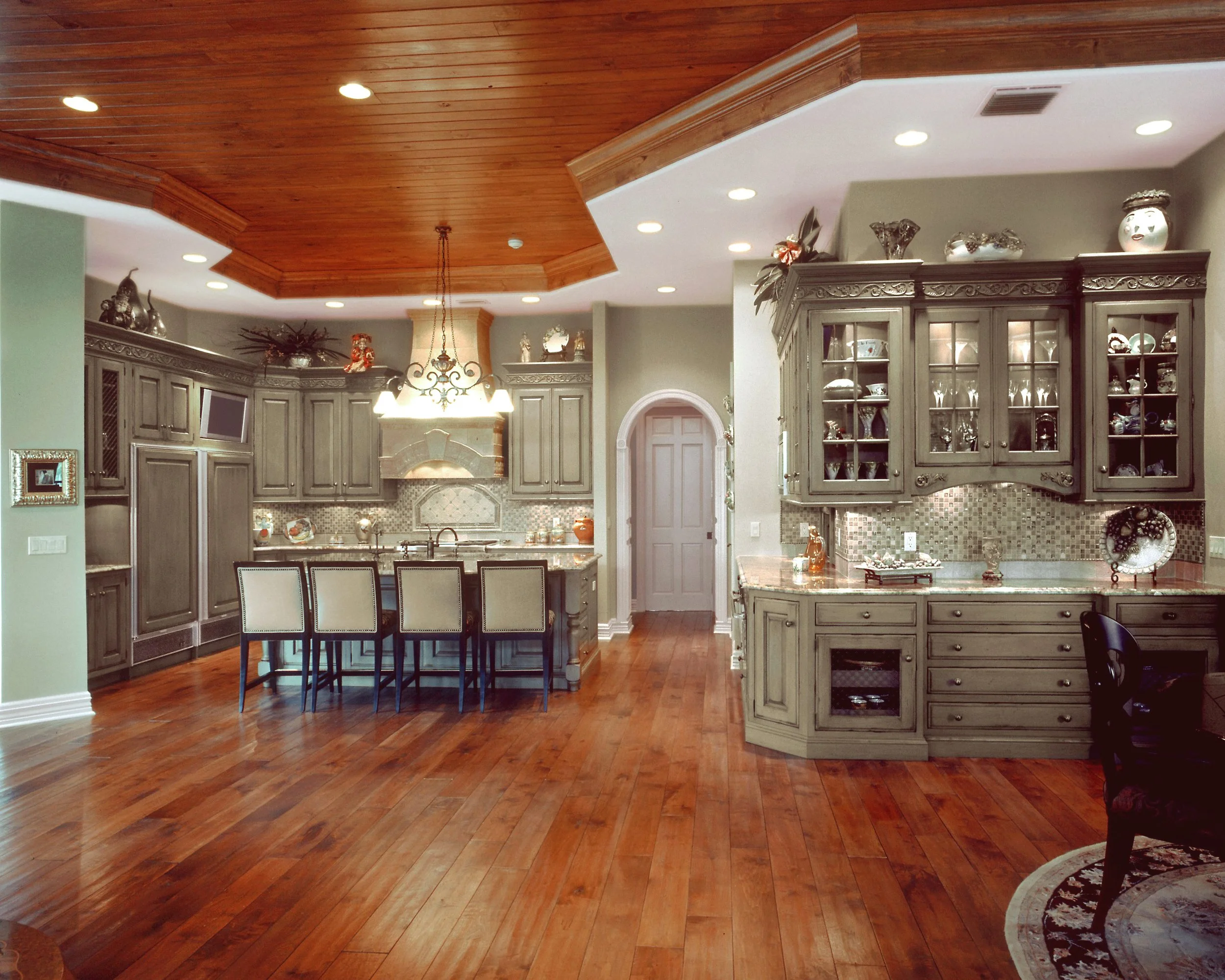 Spacious kitchen with wooden flooring, gray cabinetry, and a wooden ceiling. Features include a kitchen island with four chairs, a chandelier, a built-in oven, and decorative items on top of cabinets.