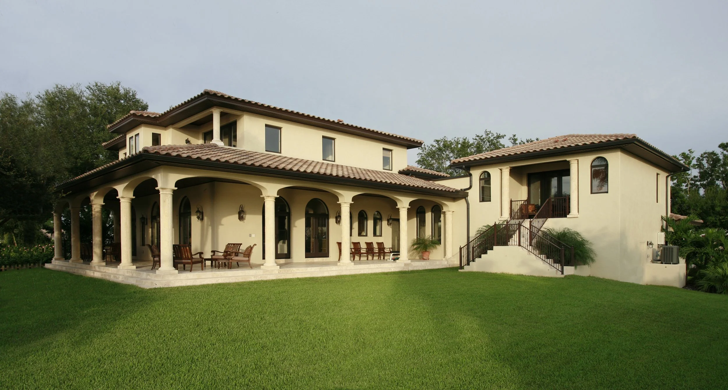 A large two-story Mediterranean-style house with a beige stucco exterior, red tile roof, and a spacious covered patio with columns, sitting on a well-manicured lawn.