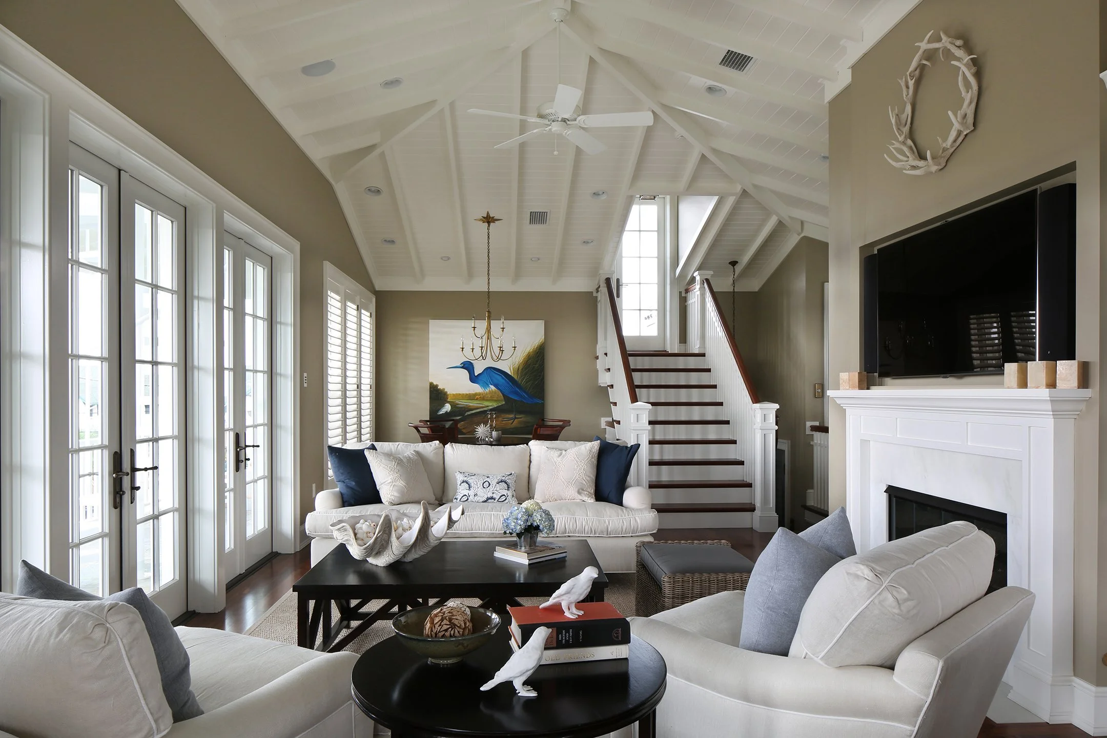 Living room with cream-colored sofa and armchair, black coffee table, and a fireplace with a large TV above. French doors on the left, stairs with white railings on the right, artwork of a blue heron behind the sofa, and a white wreath hanging on the