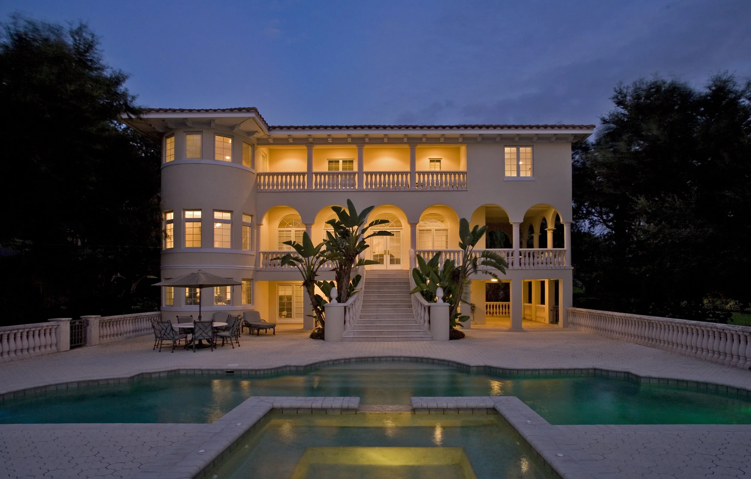 A luxurious three-story house with a white exterior illuminated at dusk, featuring arched balconies, a staircase leading to the entrance, and a swimming pool in the foreground with patio furniture and an umbrella.