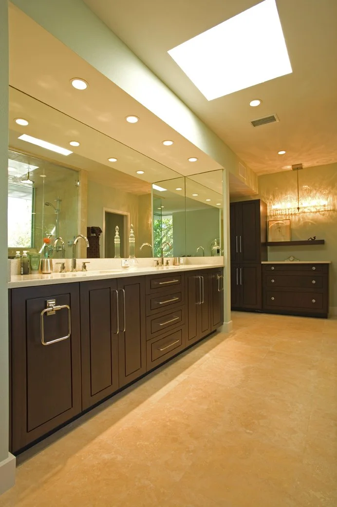 A spacious bathroom with a double vanity, large mirror, and multiple sinks. There are brown cabinets, a beige tiled floor, and a large skylight letting in natural light. A walk-in shower is visible in the background.