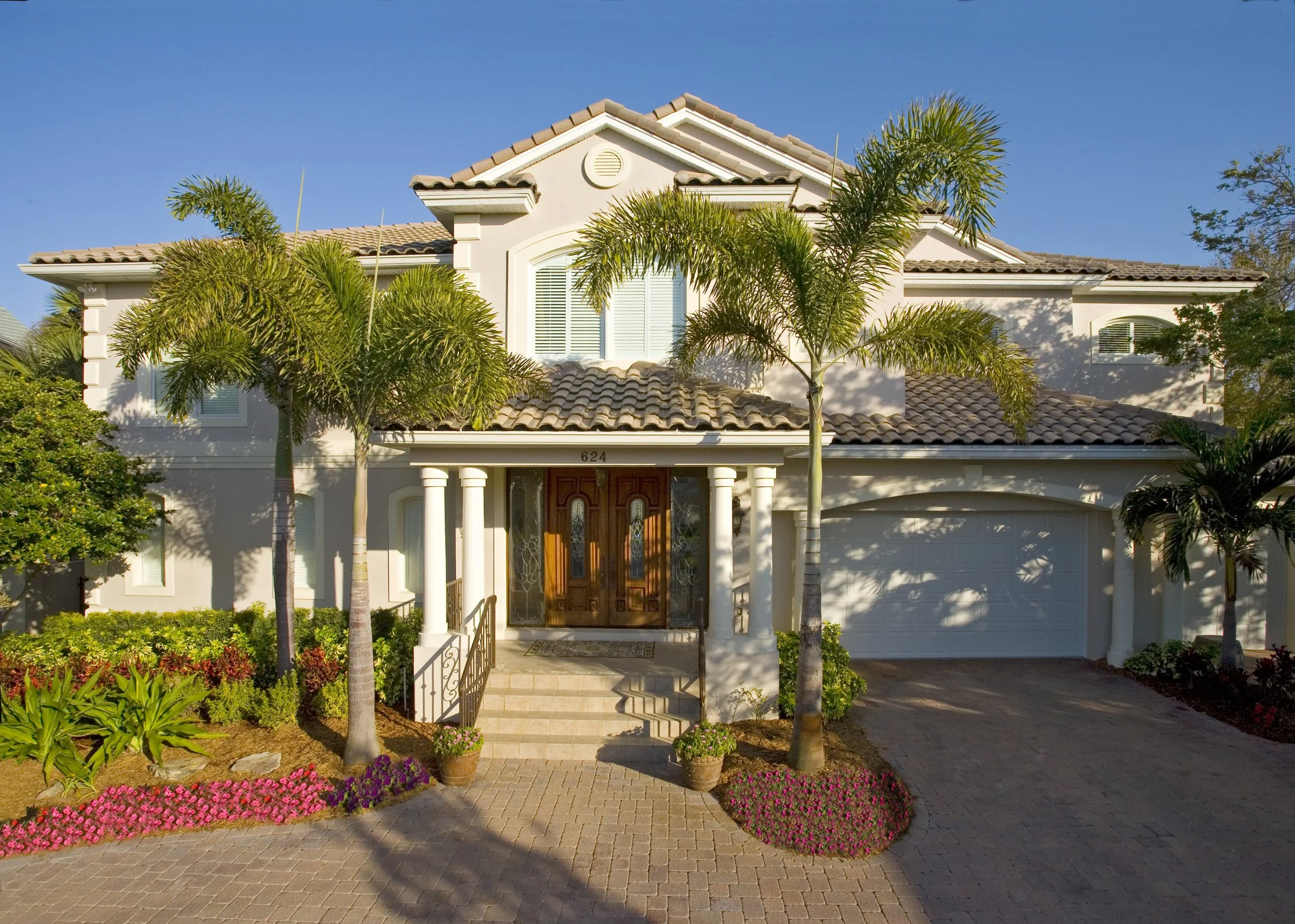 Two-story house with a tiled roof, white exterior, and a wooden front door, surrounded by palm trees and colorful flower beds, under a clear blue sky.