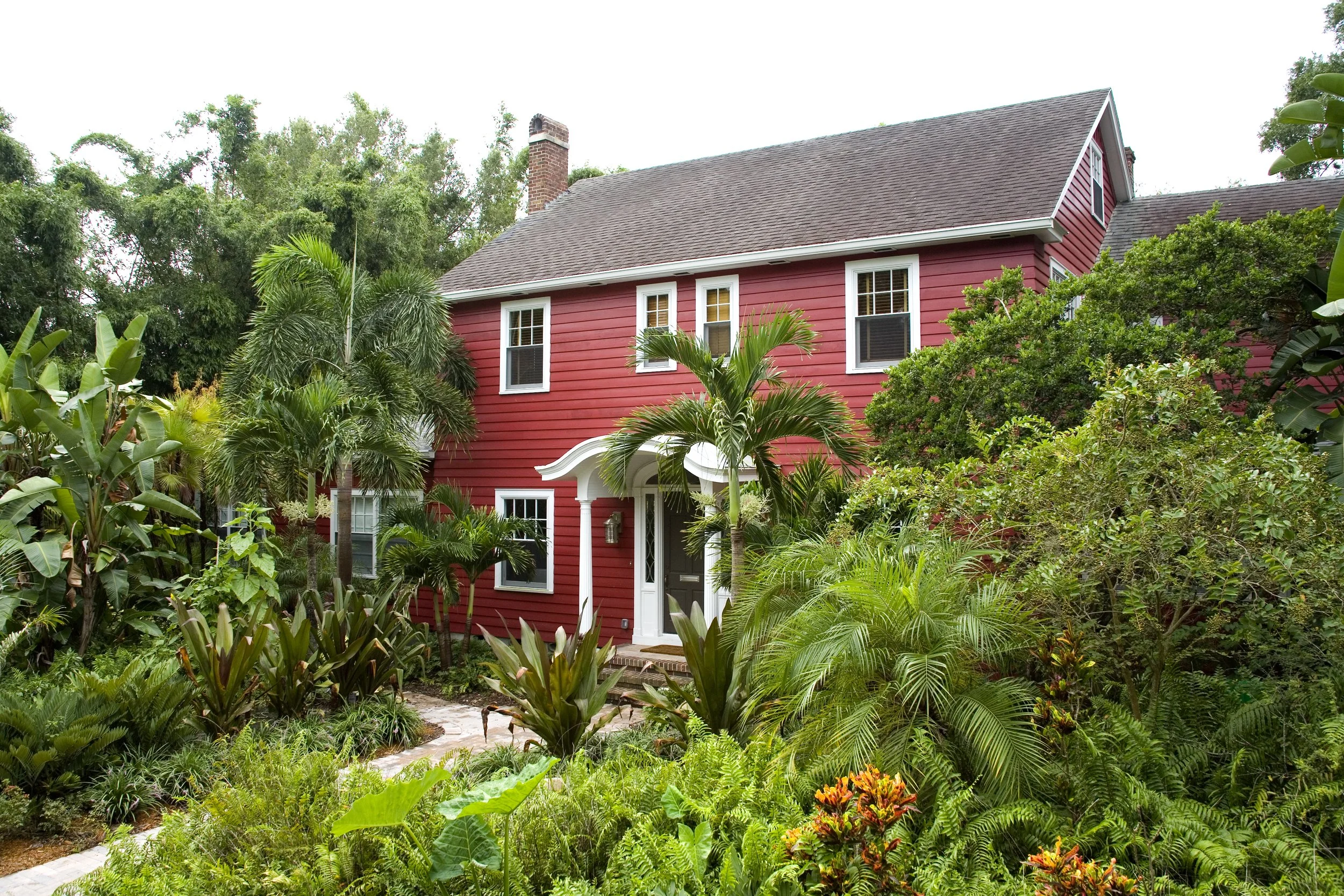 Red two-story house surrounded by lush green tropical plants and trees, with a stone pathway leading to the front door.