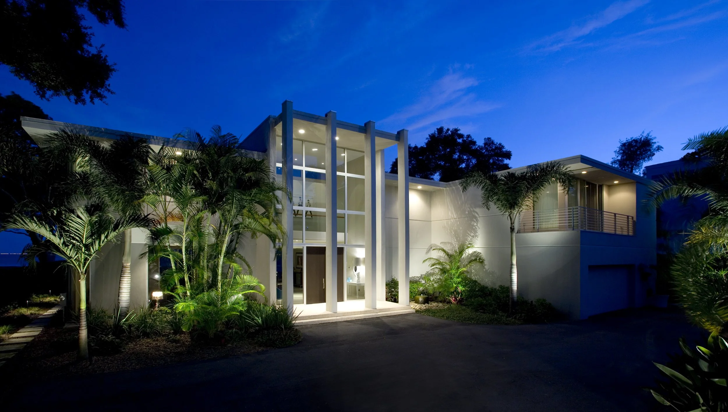 Modern house at night with well-lit entrance and surrounding lush tropical plants.