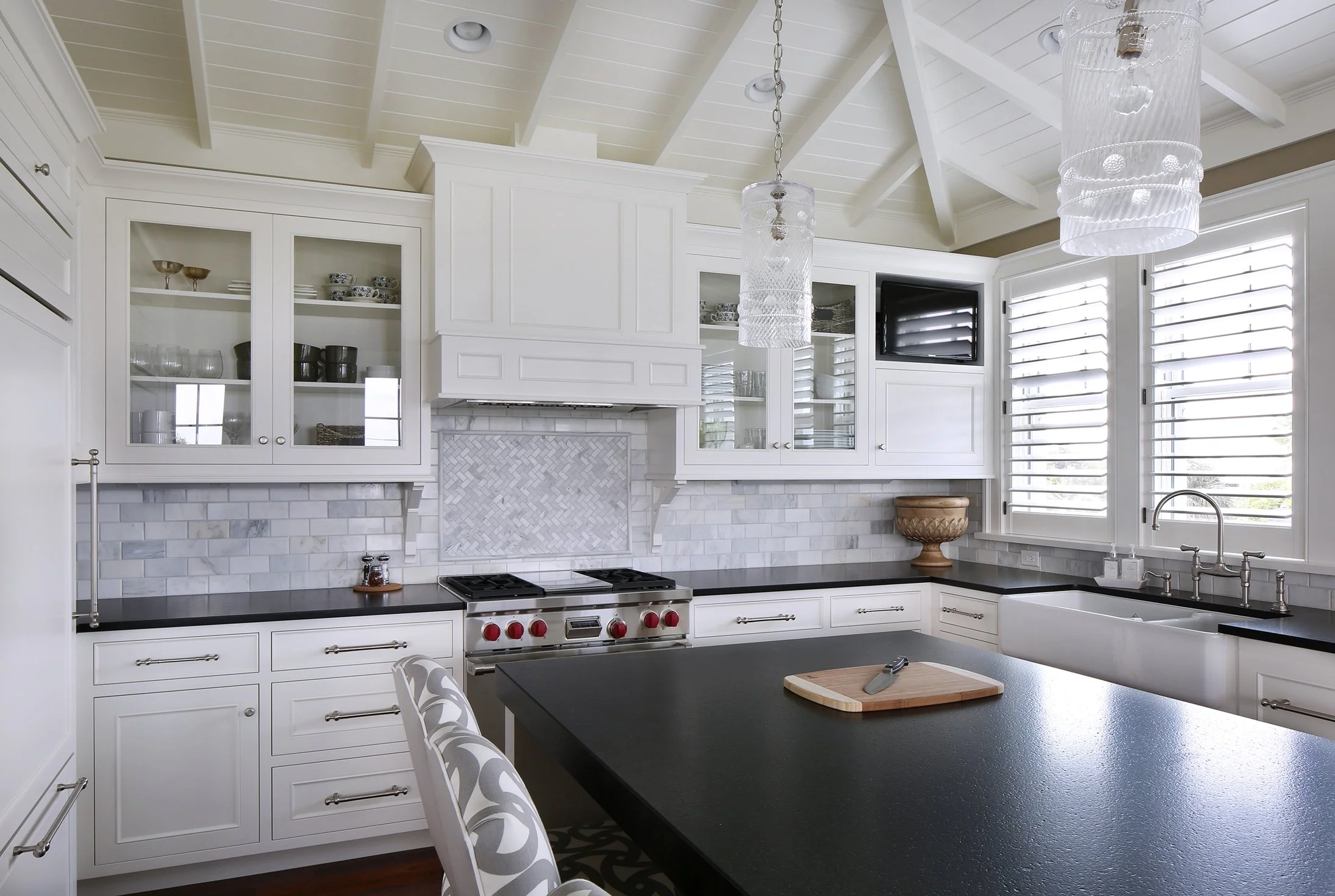 Modern white kitchen with black countertops, glass-front cabinets, stainless steel stove, and large windows with plantation shutters.