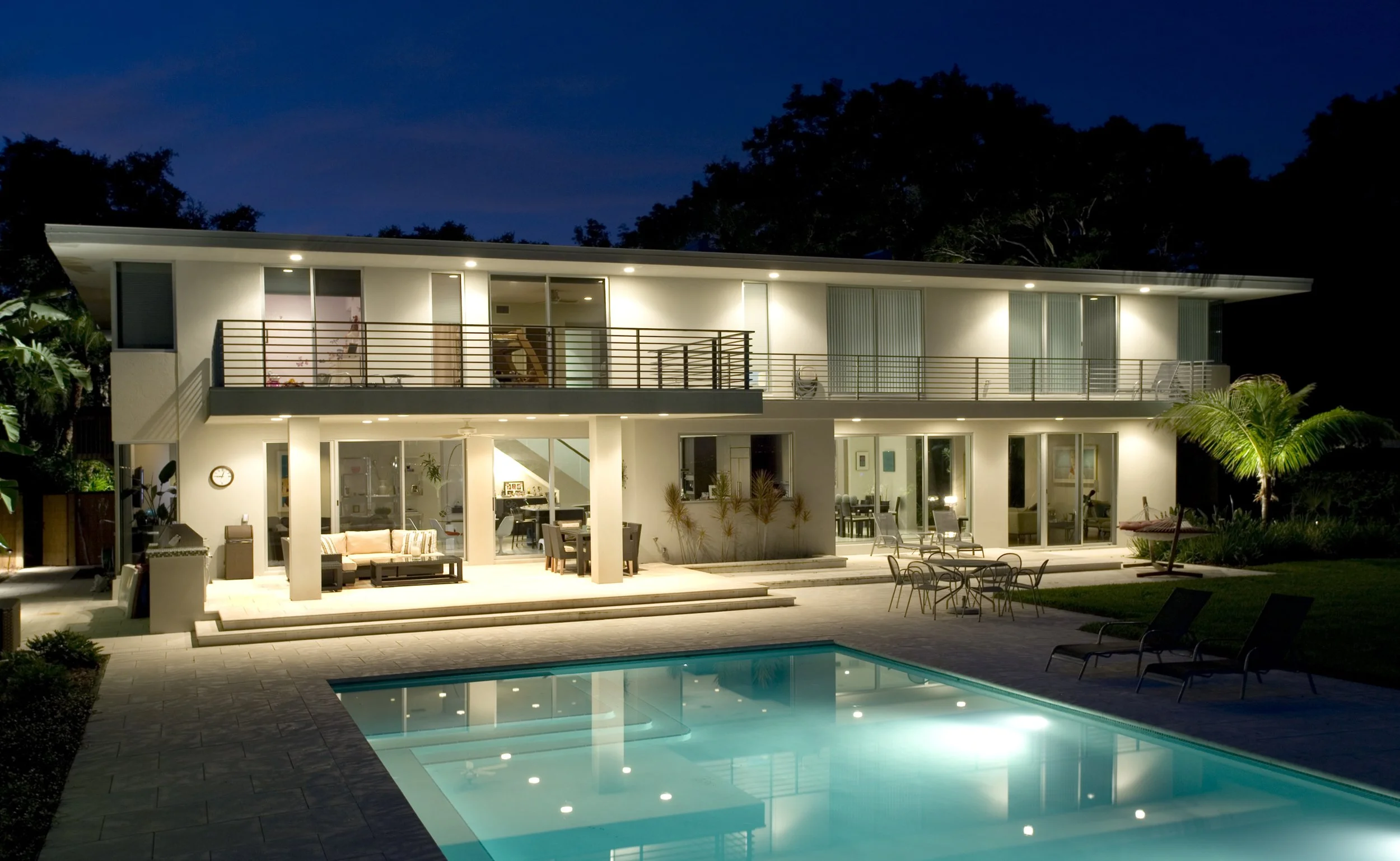 Night view of a modern two-story house with well-lit outdoor area, swimming pool, patio furniture, and surrounding greenery.