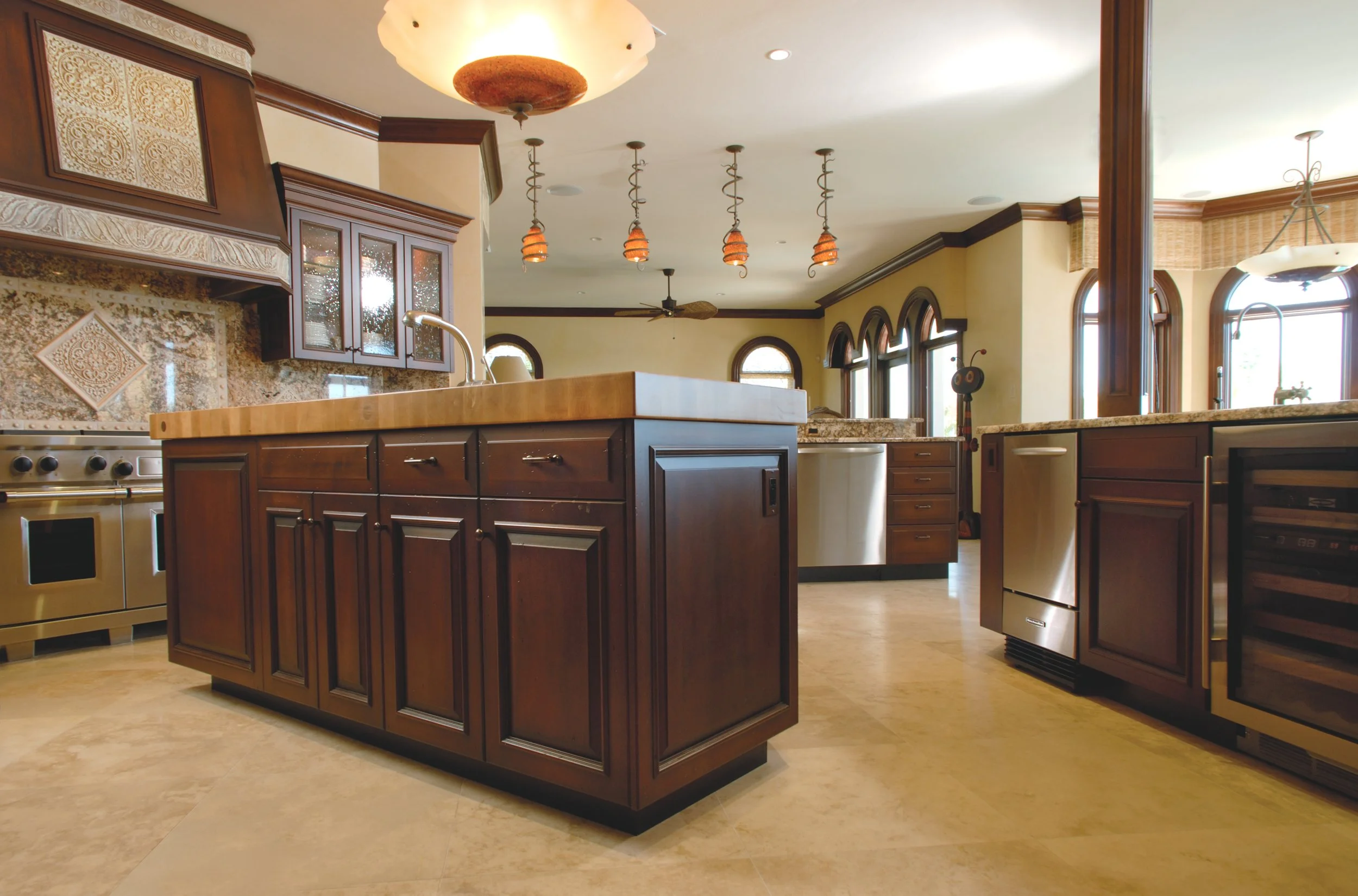 Kitchen with dark wood cabinets, beige countertops, tiled backsplash, stainless steel appliances, and multiple arched windows letting in natural light.