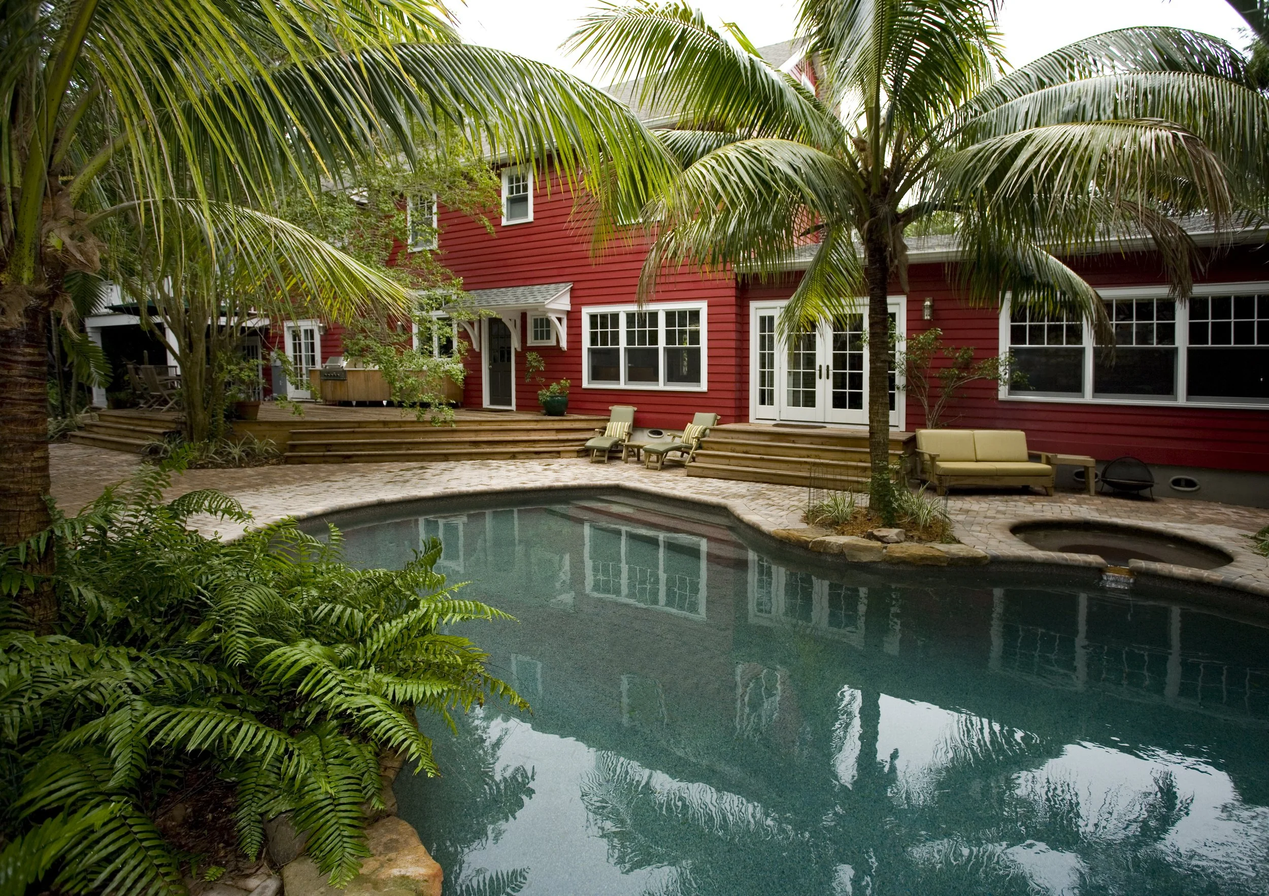 Backyard with a swimming pool, surrounded by palm trees, a red house with white trim, wooden steps, patio furniture, and a small hot tub.