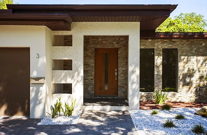 Modern house with a wooden front door, brick and stucco exterior, and large windows, surrounded by landscaped yard with white gravel and small plants.