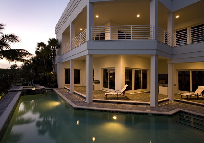 Modern two-story house with balcony, nighttime, outdoor swimming pool, lounge chairs, and palm trees.