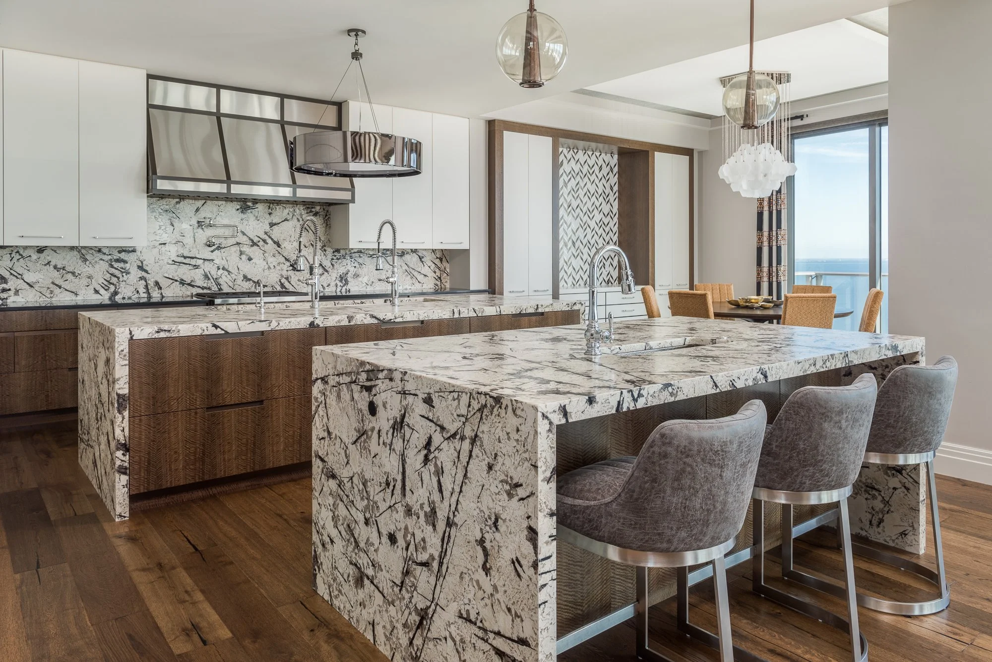 Modern kitchen with large marble island, white cabinetry, and a dining area with a view of the ocean through sliding glass doors.