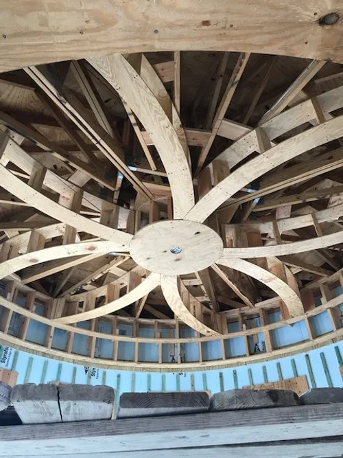 Interior view of a circular wooden structure under construction, with curved wooden beams radiating from a central hub, and an unfinished framework around the top edge.