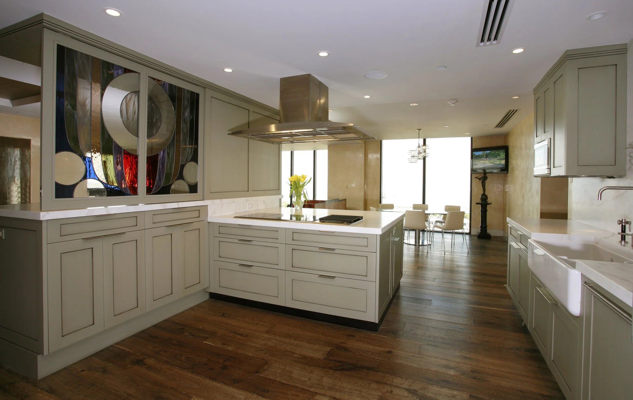 Modern kitchen with beige cabinets, white countertops, wooden floors, and an island with a stovetop, overhanging stainless steel range hood, and a vase of yellow flowers. Dining area with a glass table, chairs, and large windows in the background.
