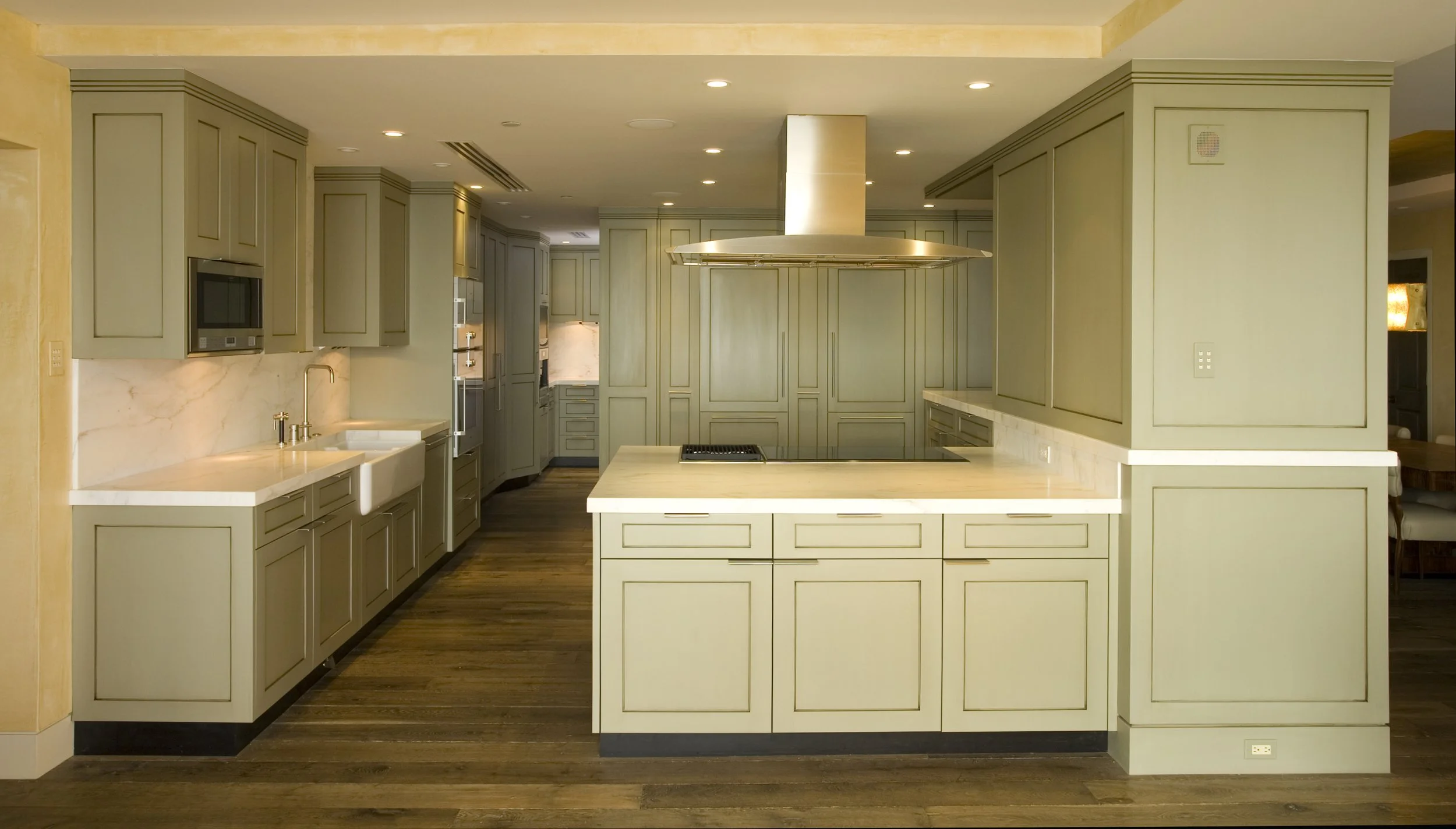 A spacious, modern kitchen with green cabinetry, white countertops, a central island with a cooktop, and a wooden floor.