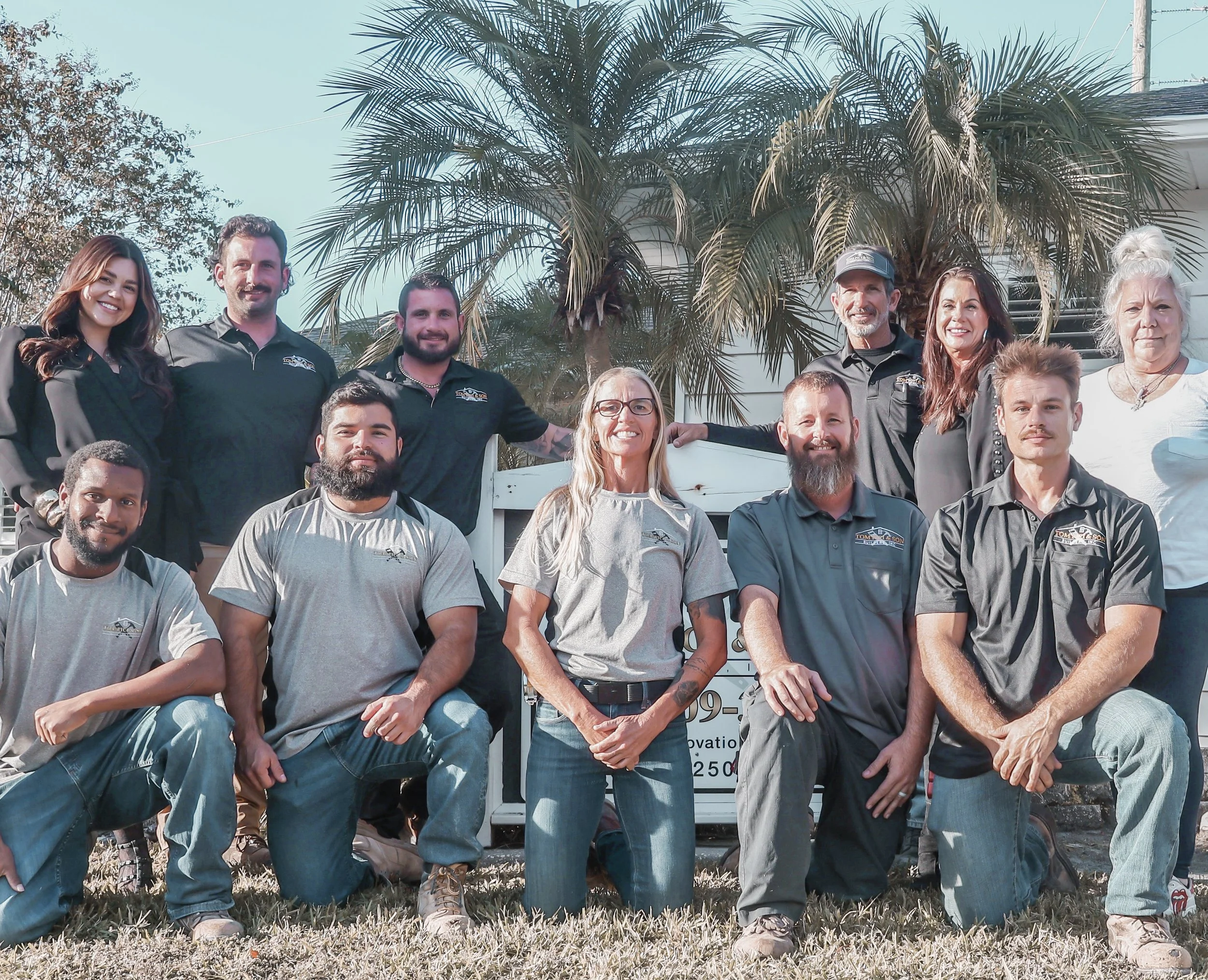 A group of eleven people, including men and women of varying ages, posing outdoors in front of a white fence and palm trees. They are casually dressed, some wearing company shirts. The photo appears to be a team or workgroup gathering on a sunny day.