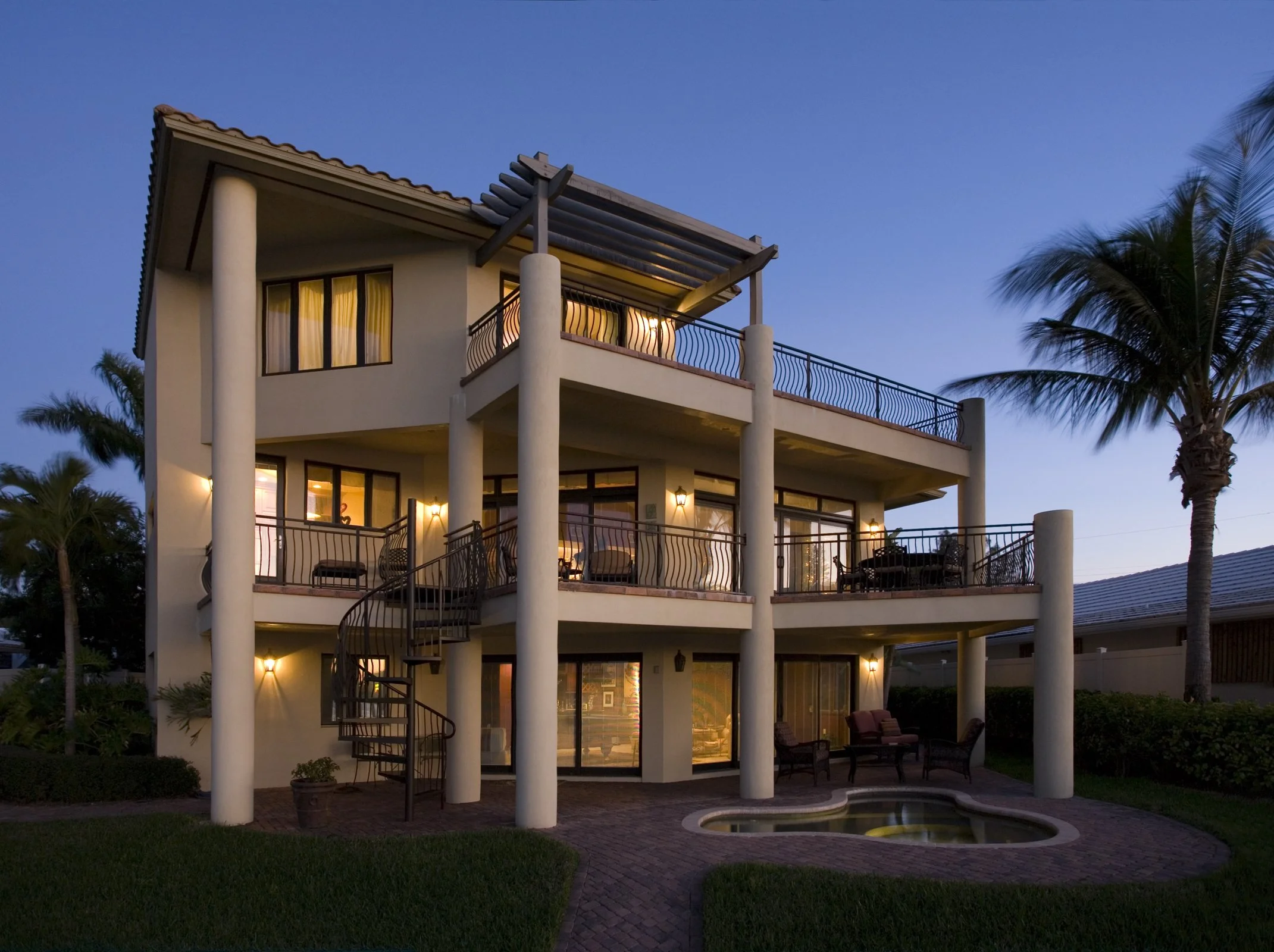 A three-story modern house with balconies on each level, large windows, and outdoor seating areas in a tropical setting with palm trees, a small pool, and evening lighting.