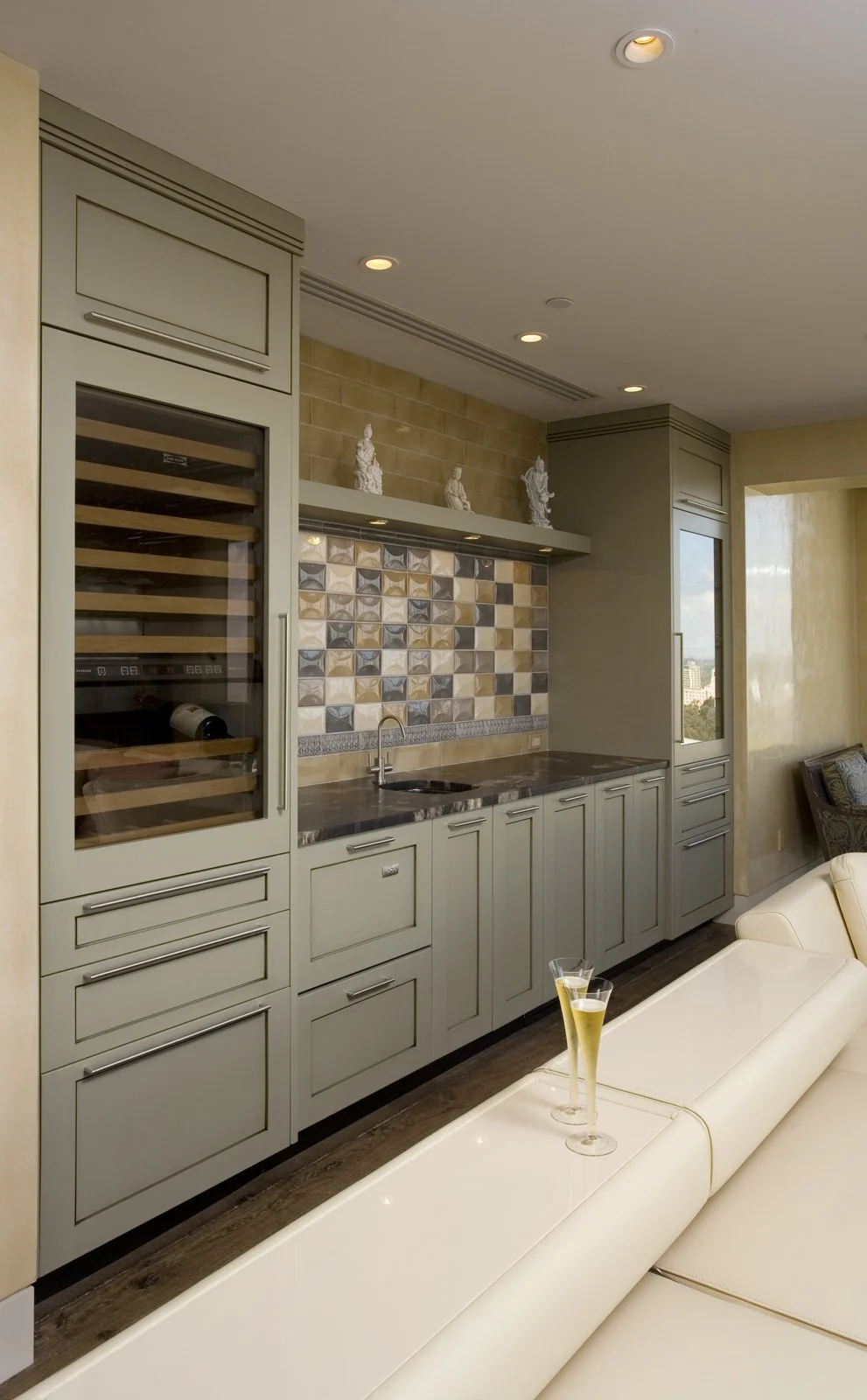 Modern kitchen with cream-colored cabinets, a decorative tile backsplash, a small sink, and two glasses of sparkling wine on a white sofa.