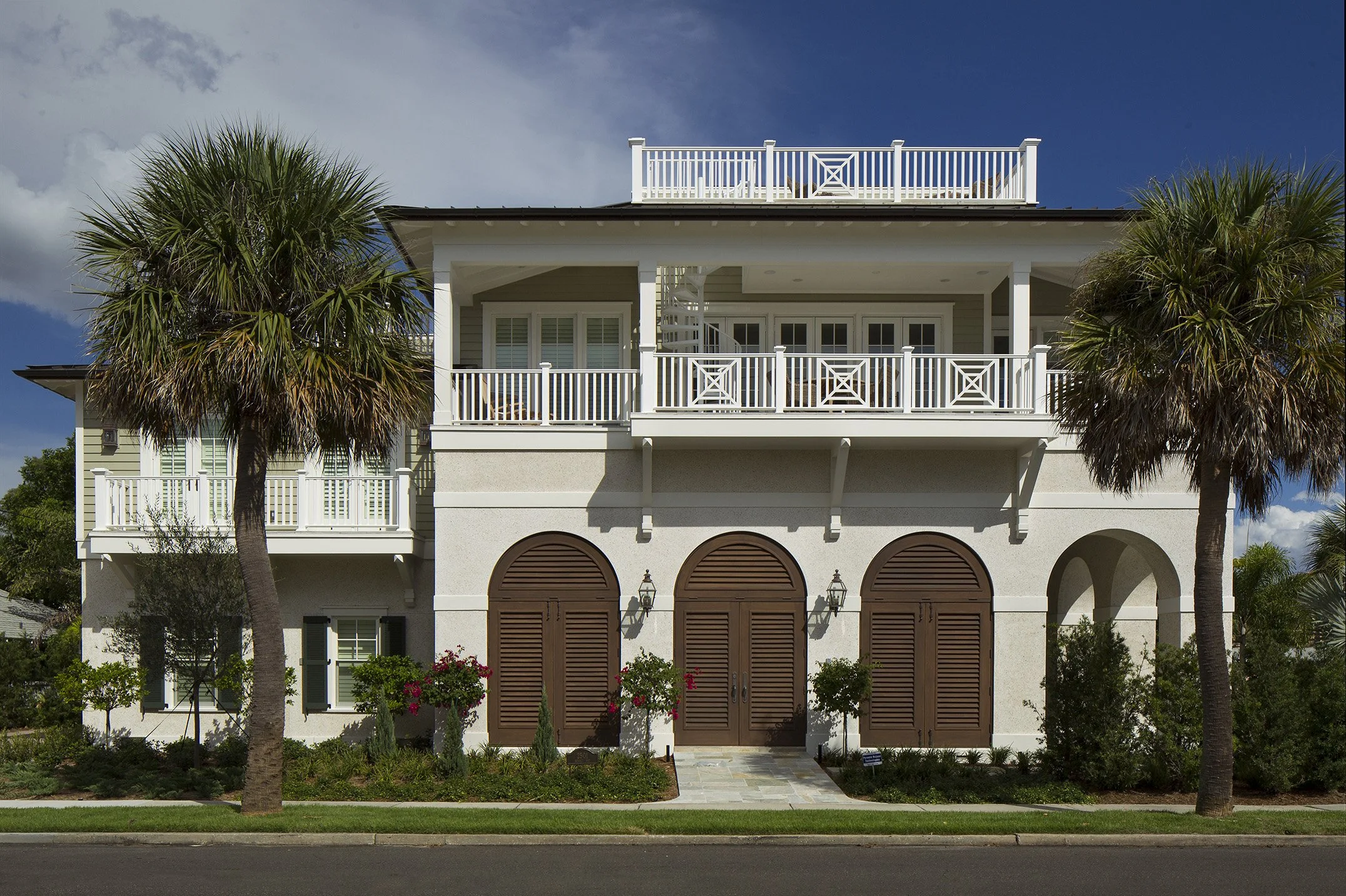 A large, white, multi-story house with brown shutters, multiple balconies, and surrounded by palm trees and landscaped greenery under a partly cloudy sky.