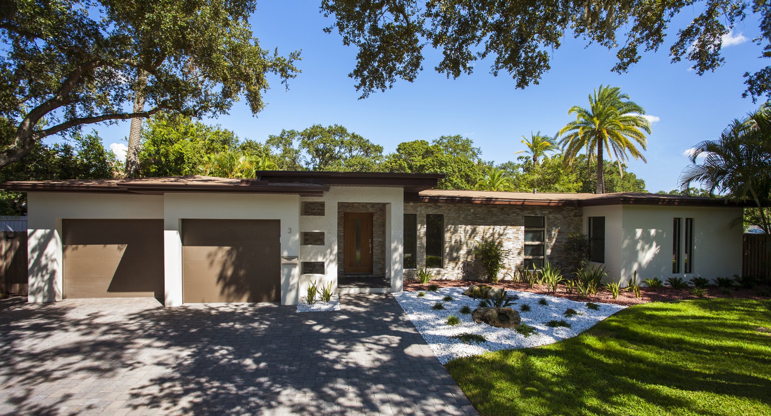 Modern single-story house with a landscaped front yard, double garage, and a tree casting shadows on the driveway.