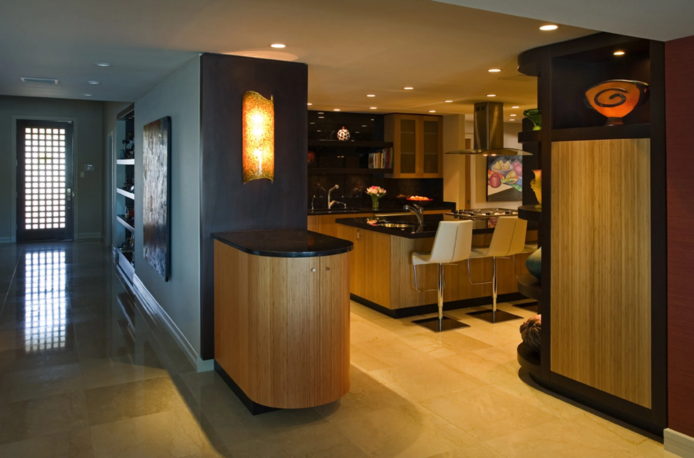 Interior view of a modern kitchen with wooden cabinets, black countertops, and white bar stools around a kitchen island, with built-in shelves and decorative art.