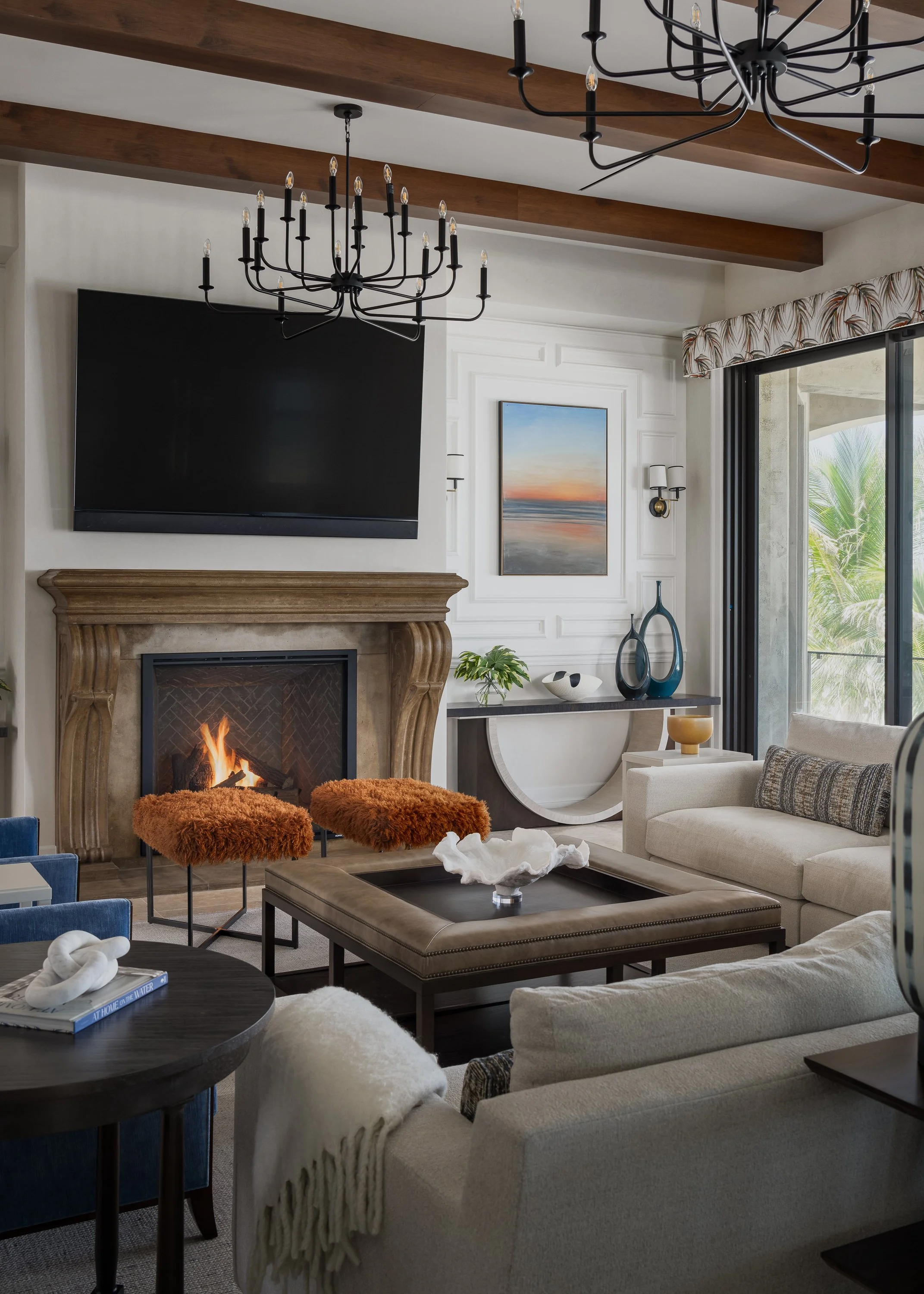Living room with a fireplace, wall-mounted TV, two orange furry stools, a beige sofa, a black coffee table, a sideboard with blue vases, and sliding glass doors showing trees outside.