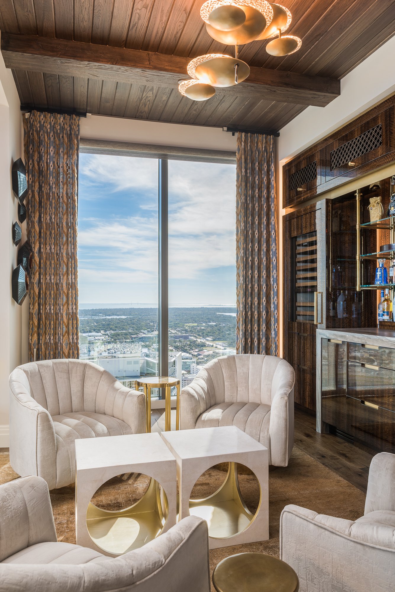 Living room with two white velvet armchairs, a small gold side table, a marble coffee table, and large floor-to-ceiling window with a city view. There are patterned curtains, a modern ceiling light fixture, and wood accents.