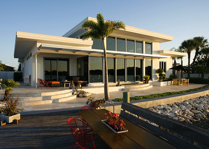 Modern two-story house with large glass windows, surrounded by palm trees, with outdoor seating and a rocky landscape.