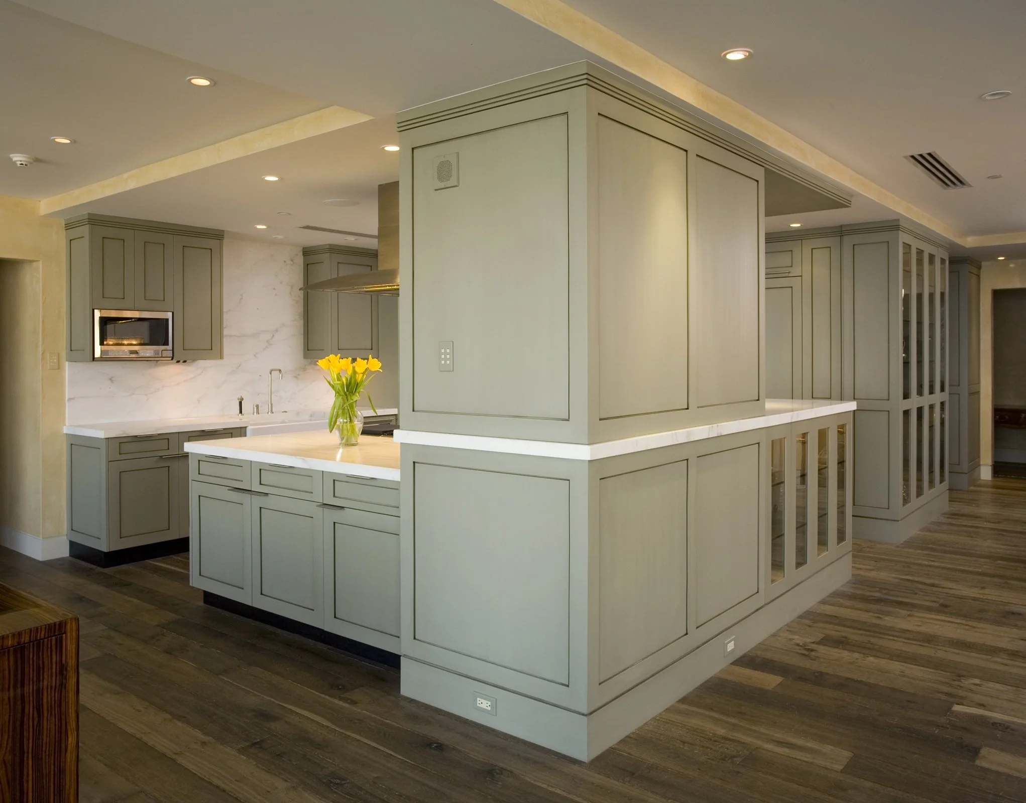 A modern kitchen with green cabinetry, white marble backsplash, and a kitchen island with a vase of yellow flowers.