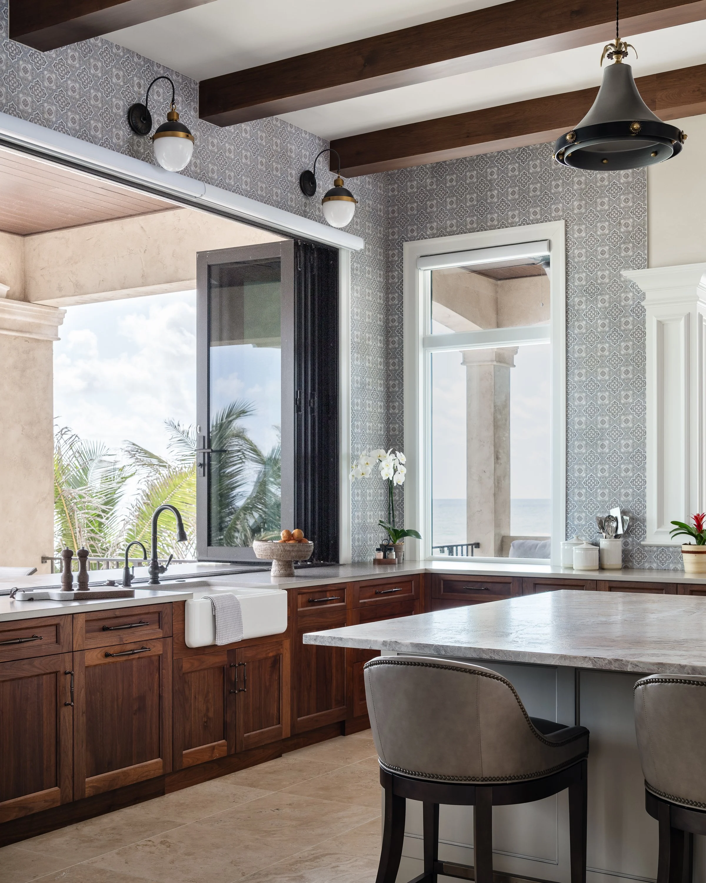 A kitchen with wooden cabinets, a white farmhouse sink, and a large window showing an outdoor view with palm trees and ocean