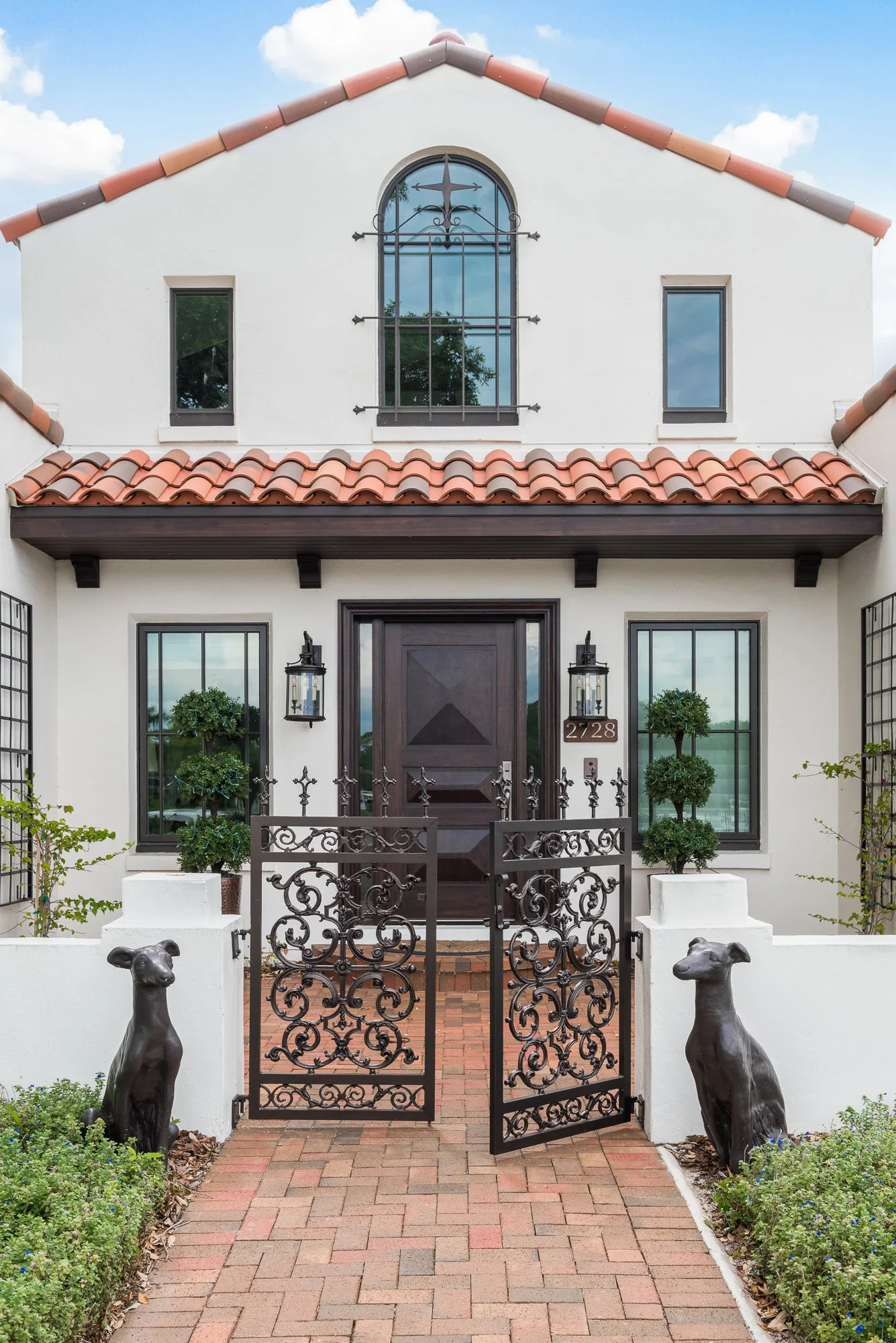 Front view of a white house with a tiled roof, iron gate, potted trees, and decorative dog statues.
