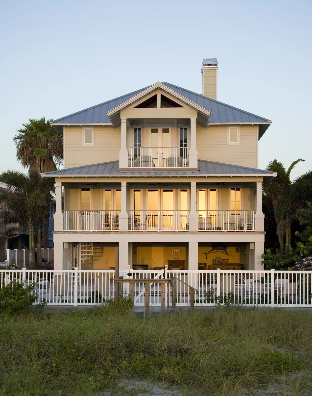 Multi-story beach house with balconies, surrounded by palm trees, fenced yard, and grassy area in front, during sunset.