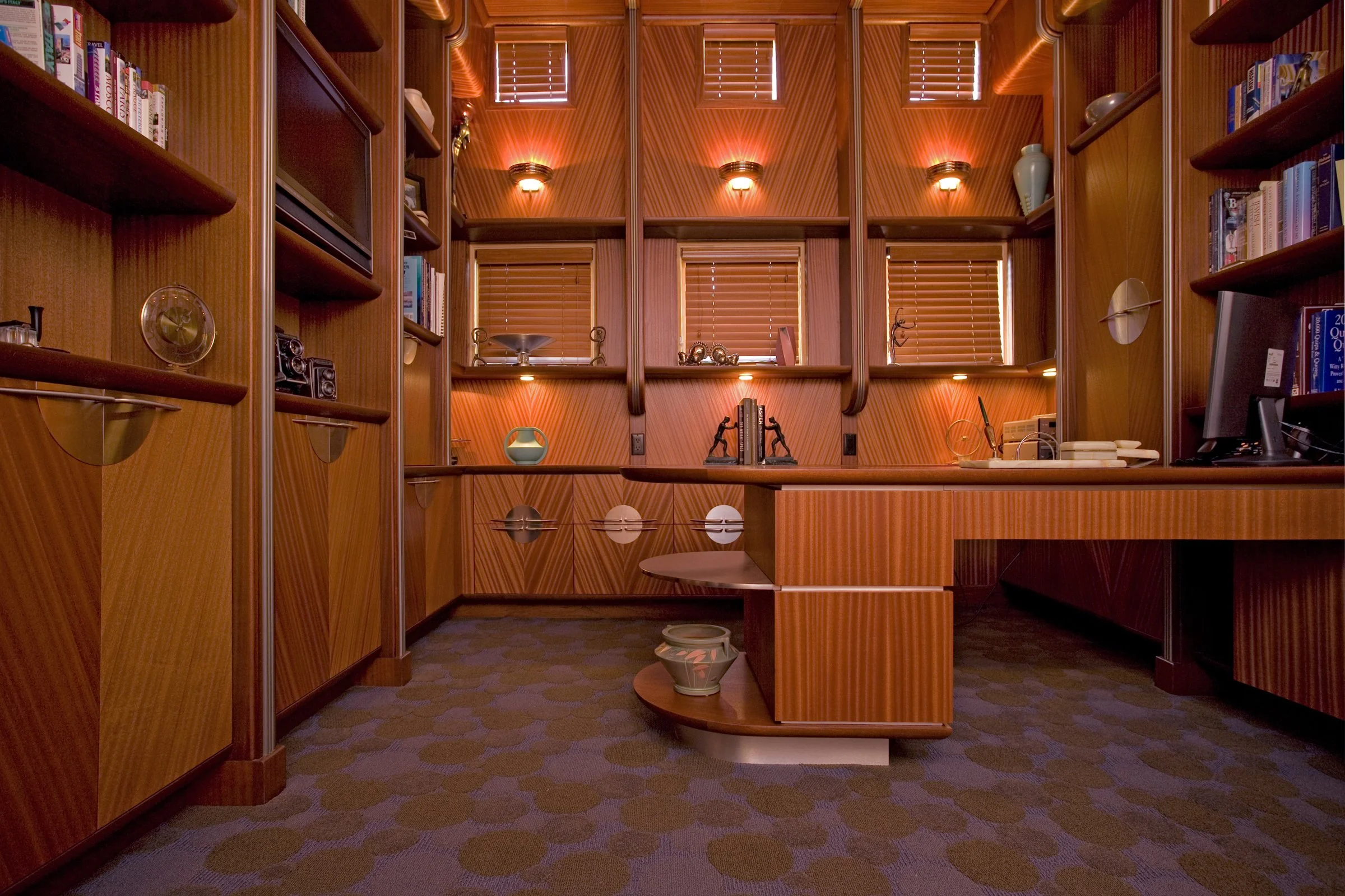 A cozy wood-paneled home office with built-in shelves, a desk, and warm lighting, containing books, decorative items, and a computer monitor.