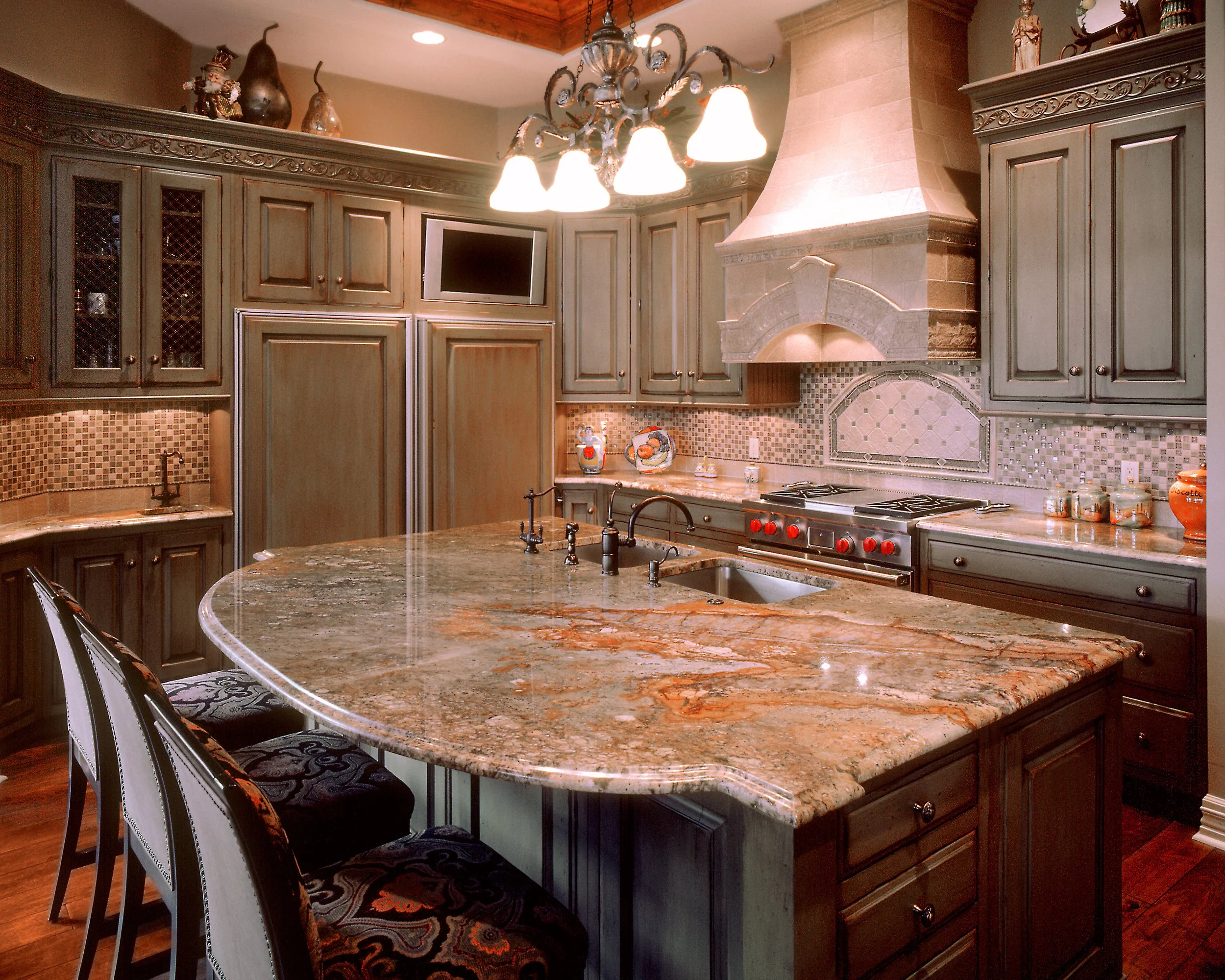 A spacious kitchen with wooden cabinets, a large granite island, a stove with red knobs, a hood, a small TV, and decorative items on top of the cabinets. There are bar stools with patterned cushions around the island, and a chandelier hanging from th