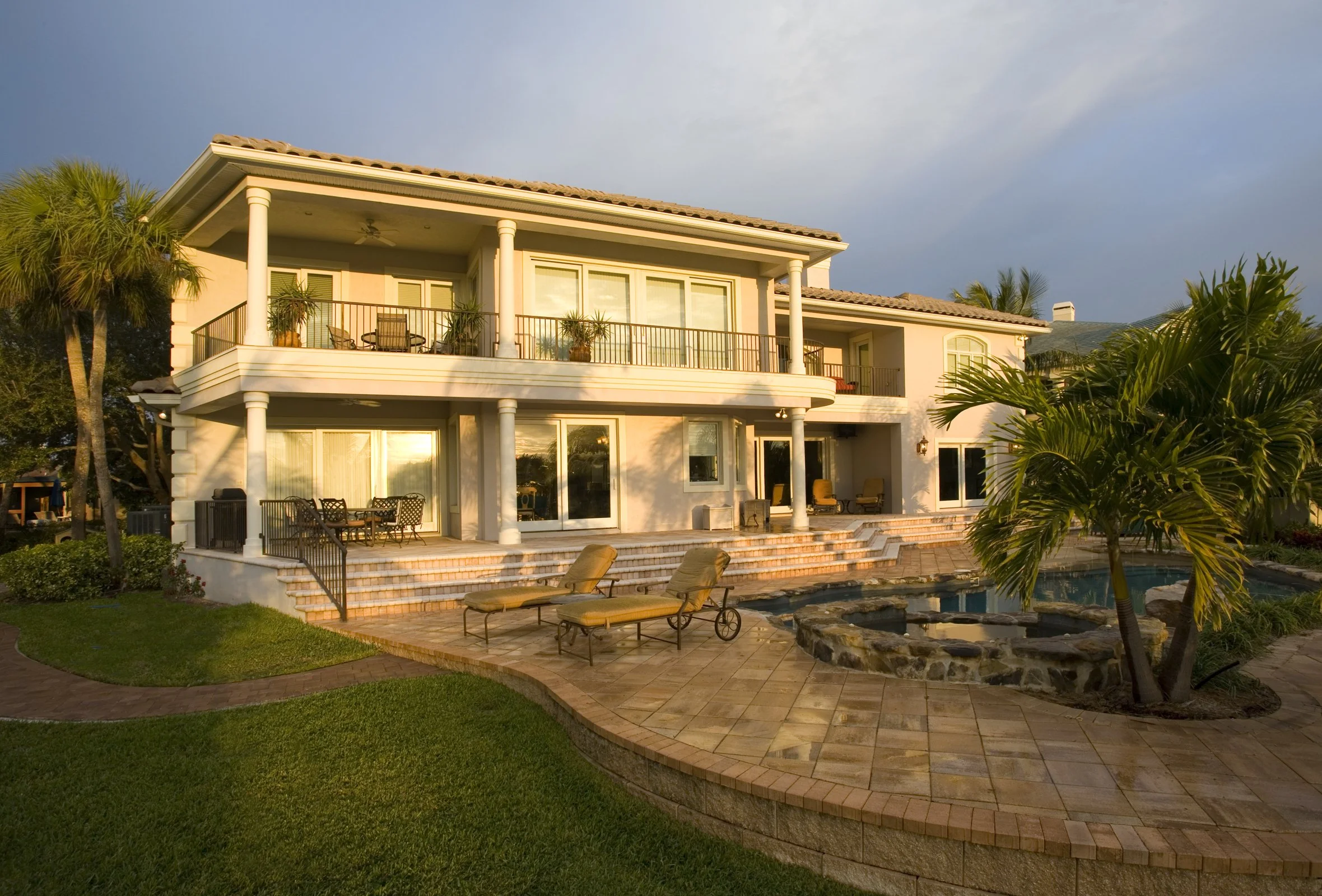 Large two-story house with balconies, outdoor furniture, pool, and palm trees at sunset