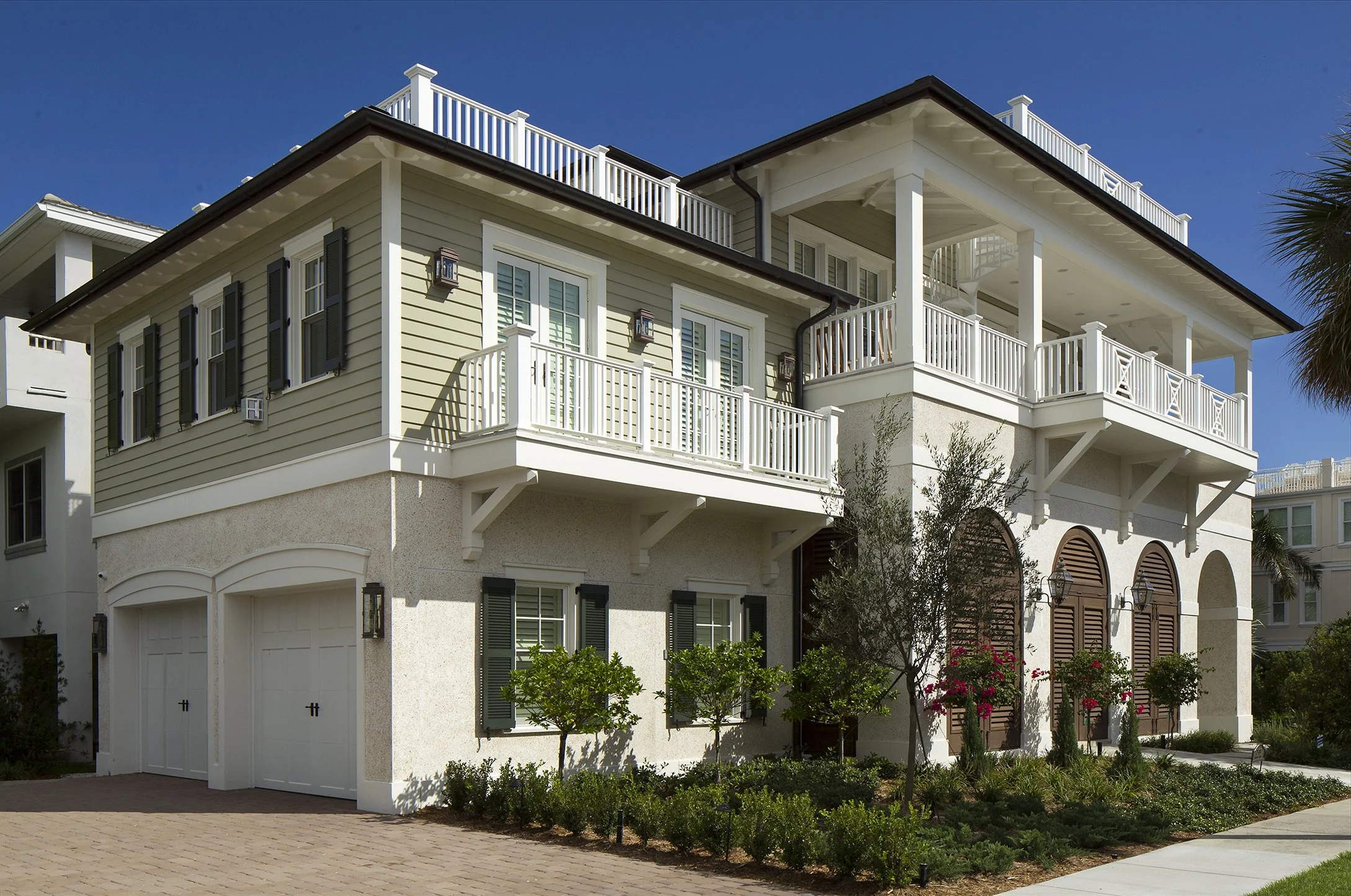 A multi-story house with beige siding, black shutters, and white trim. It features a two-car garage, a manicured front yard with small trees and shrubs, and balconies with white railings. The sky is clear and blue.