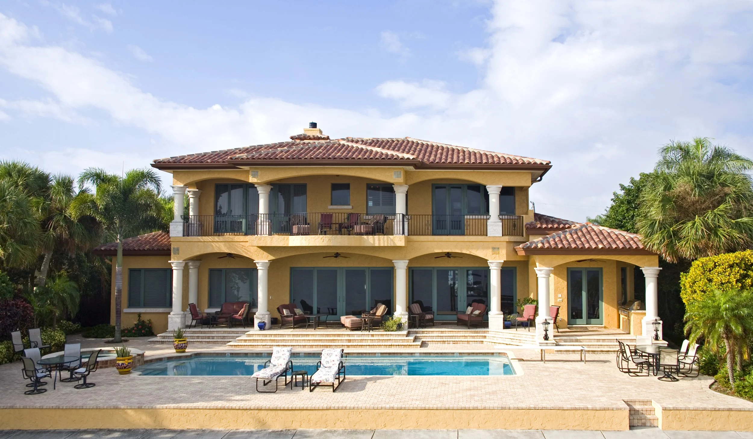 Large multi-story house with a swimming pool and outdoor seating area, surrounded by palm trees, under a partly cloudy sky.